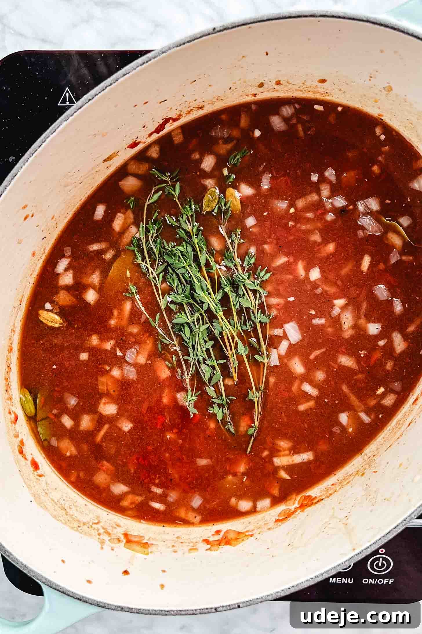 Melt-in-Your-Mouth Oven Braised Lamb Shanks 8 The simmering, aromatic braising liquid in an oval pot, featuring cooked diced onions, whole cardamom pods, a vibrant red hue from tomatoes and pomegranate, and fresh thyme sprigs, ready to welcome the lamb.