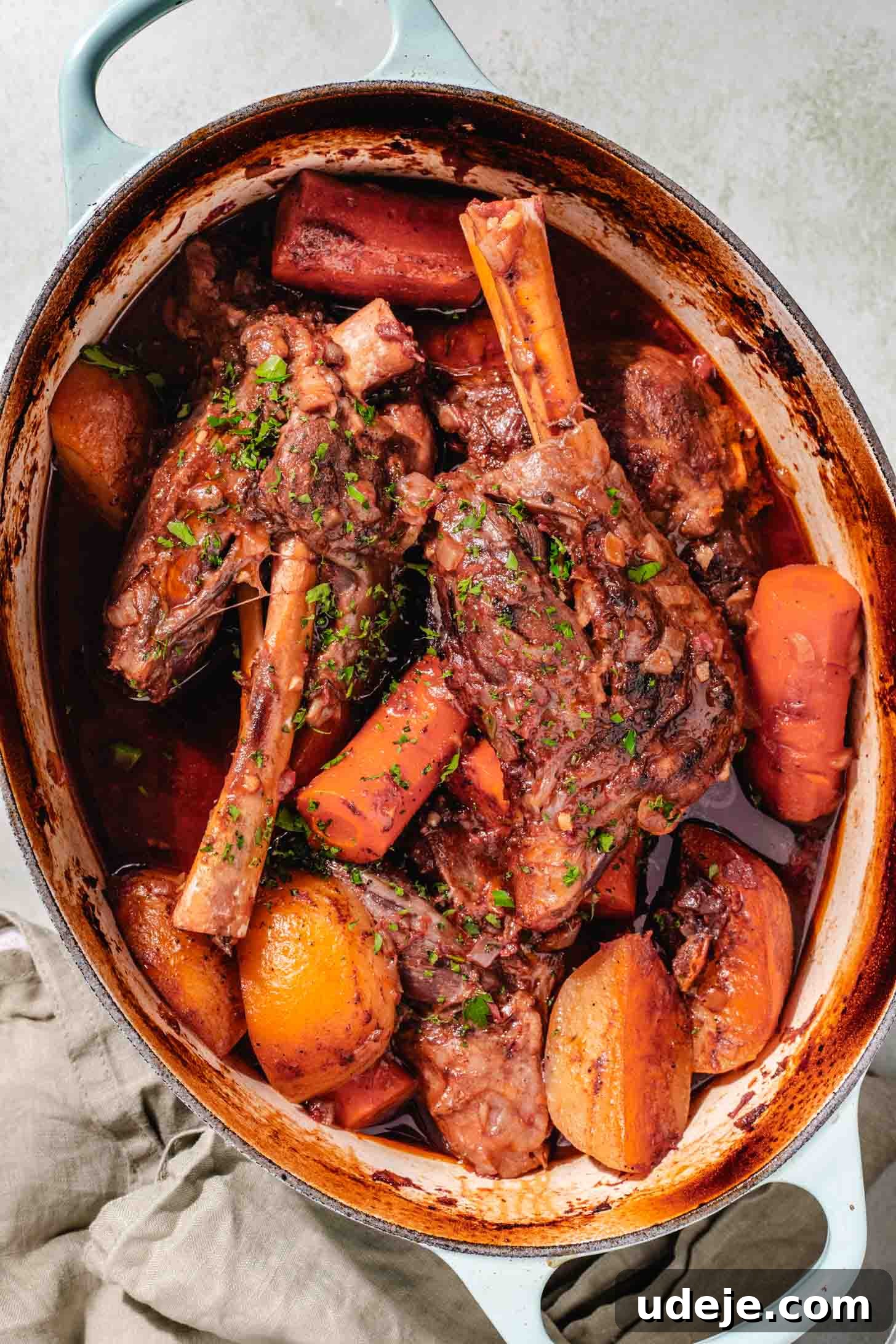 Melt-in-Your-Mouth Oven Braised Lamb Shanks 10 A large oval Dutch oven proudly displays the finished braised lamb shanks, surrounded by tender carrot pieces and potato wedges, all swimming in a rich, deeply flavored broth, ready to be served. The pot is set on a sage green background with a matching tea towel.