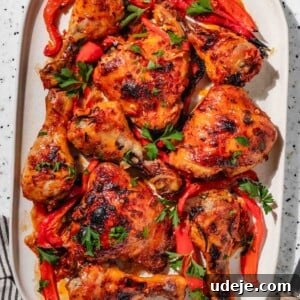 Baked red chicken with sliced red peppers on a rectangular white platter with a green glass of water, a black and white striped tea towel, a wooden handled knife and a small plate of parsley leaves on a speckled background.