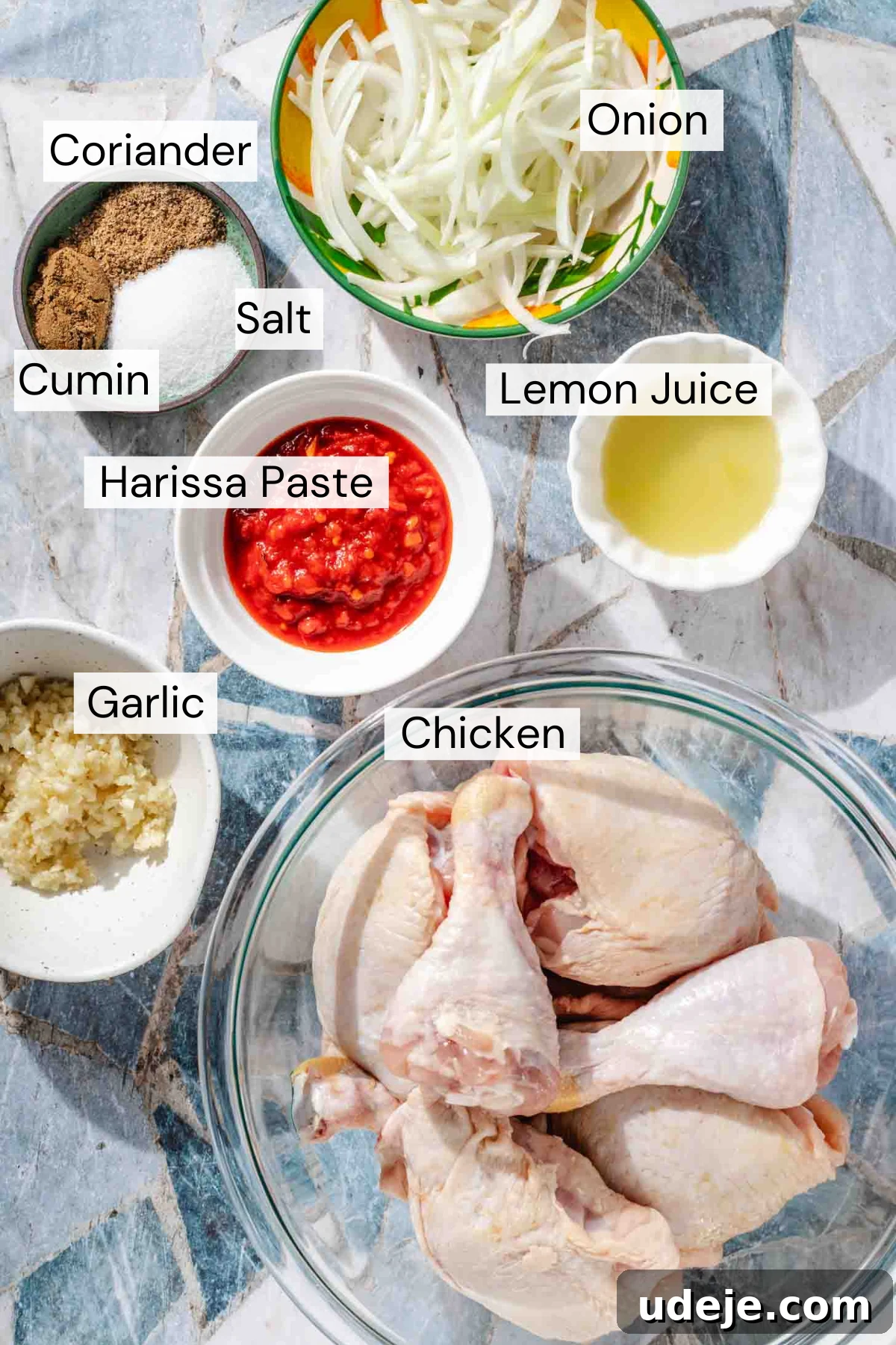 Sticky Harissa Honey Glazed Chicken 3 Ingredient shot with sliced onions in a yellow and green bowl, a small bowl with coriander, cumin and salt, a small white bowl with lemon juice, a small white bowl with red harissa paste, a small white bowl with minced garlic, a glass bowl with chicken legs and thighs on a light blue and white background.