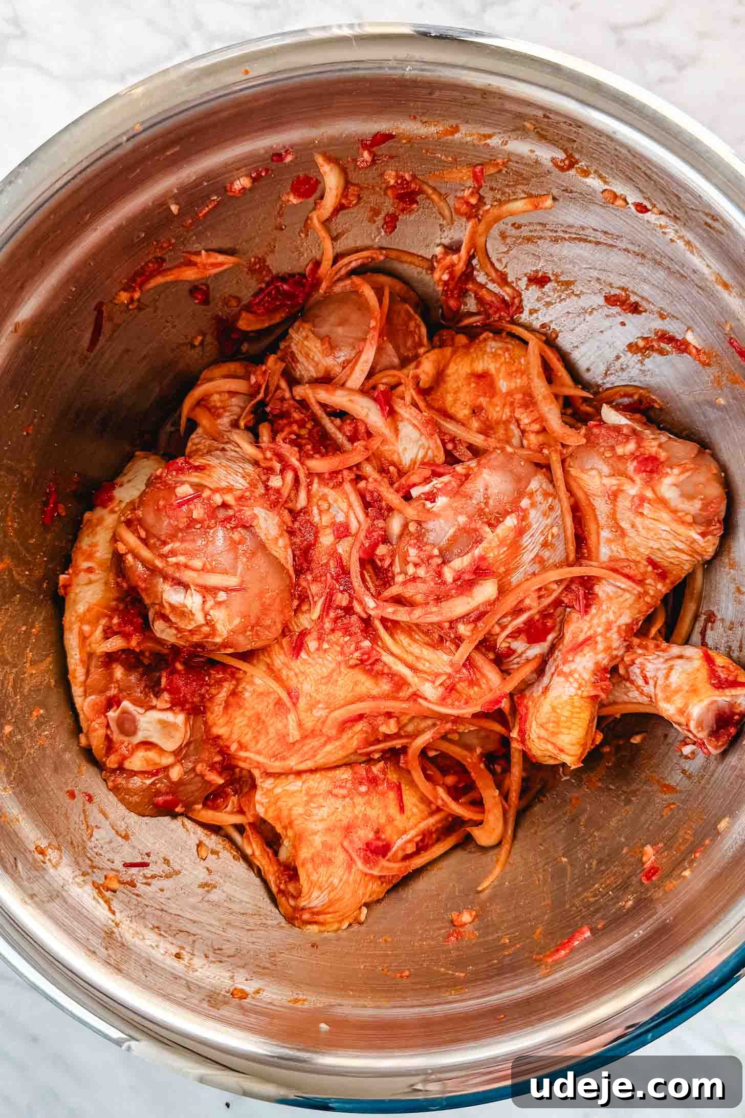 Sticky Harissa Honey Glazed Chicken 4 Chicken legs and thighs marinated in a red paste with sliced onions in a metal bowl on a marble background.