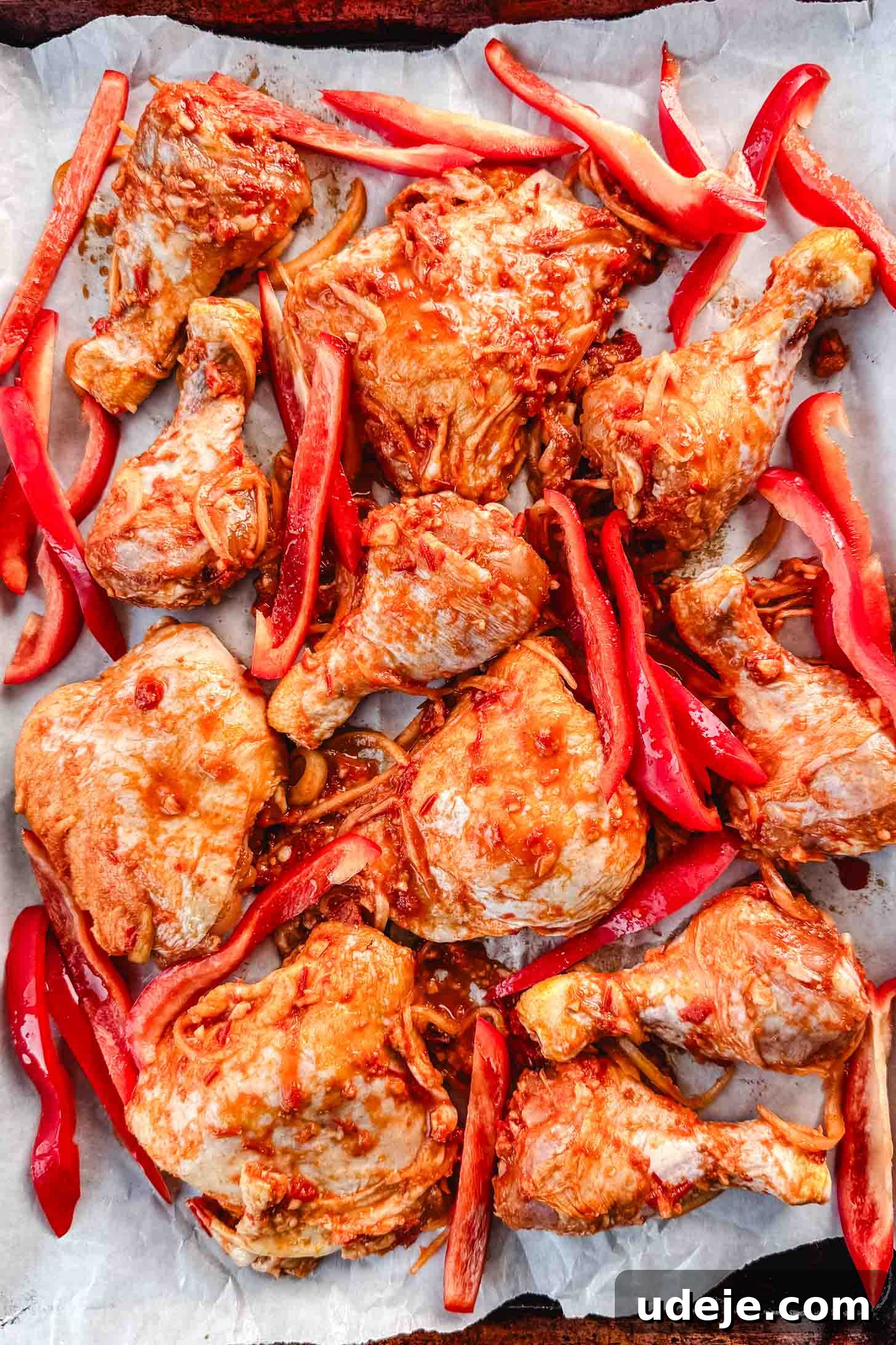 Sticky Harissa Honey Glazed Chicken 5 Chicken legs and thighs marinated in a red coloured marinade with sliced onions and sliced red peppers on a parchment lined baking sheet.