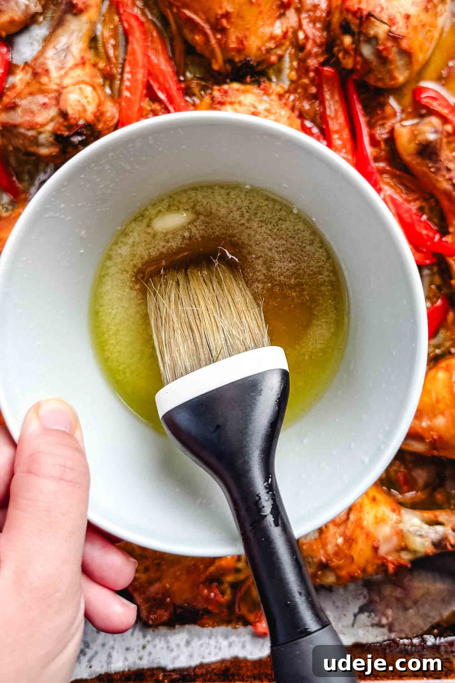 Sticky Harissa Honey Glazed Chicken 6 Honey and butter melted together in a small white bowl being held on top of baked red chicken thighs and legs and sliced red peppers with a pastry brush in the bowl.