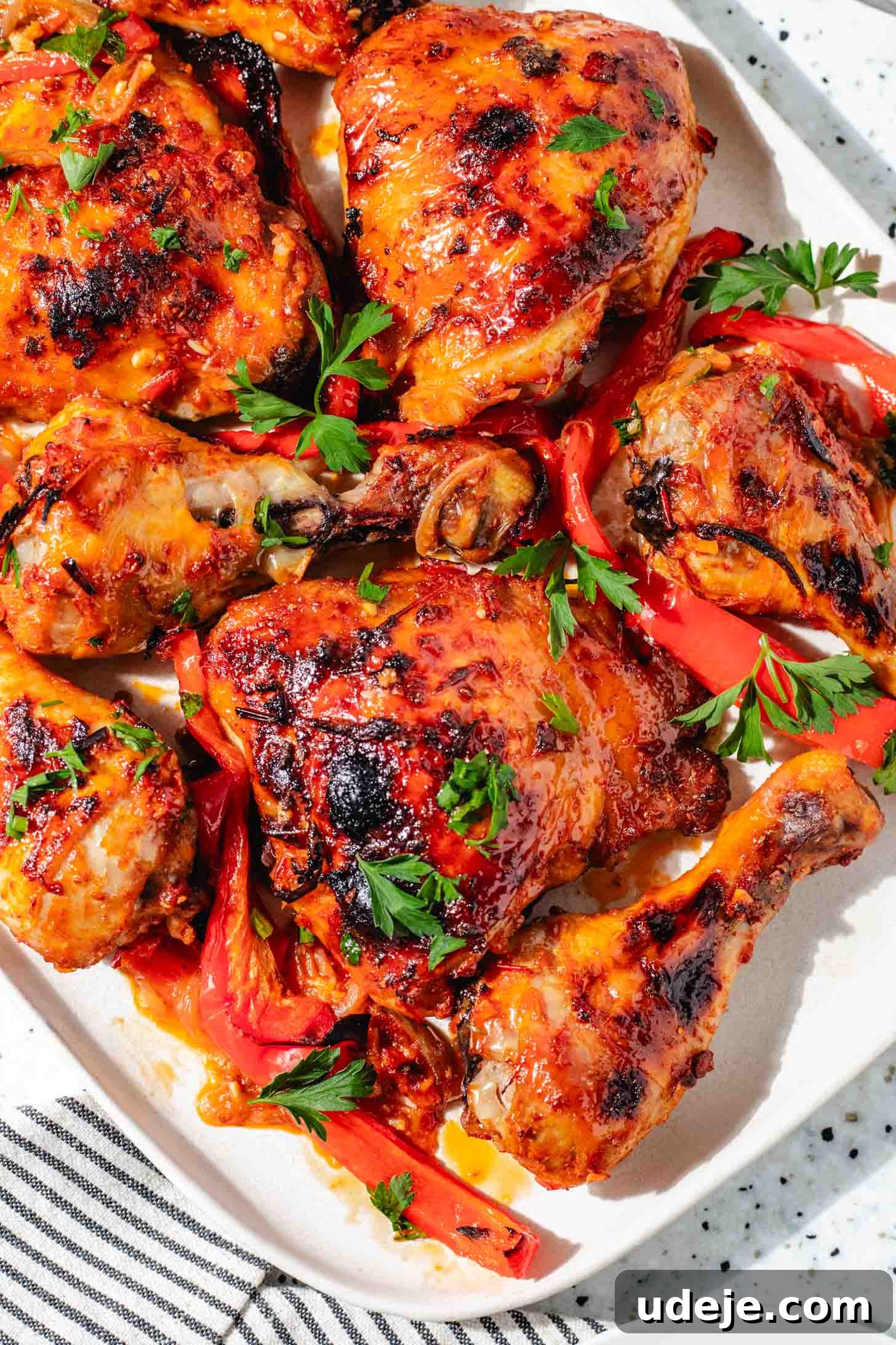 Sticky Harissa Honey Glazed Chicken 8 Baked red chicken with sliced red peppers on a rectangular white platter with a black and white striped tea towel on a speckled background.