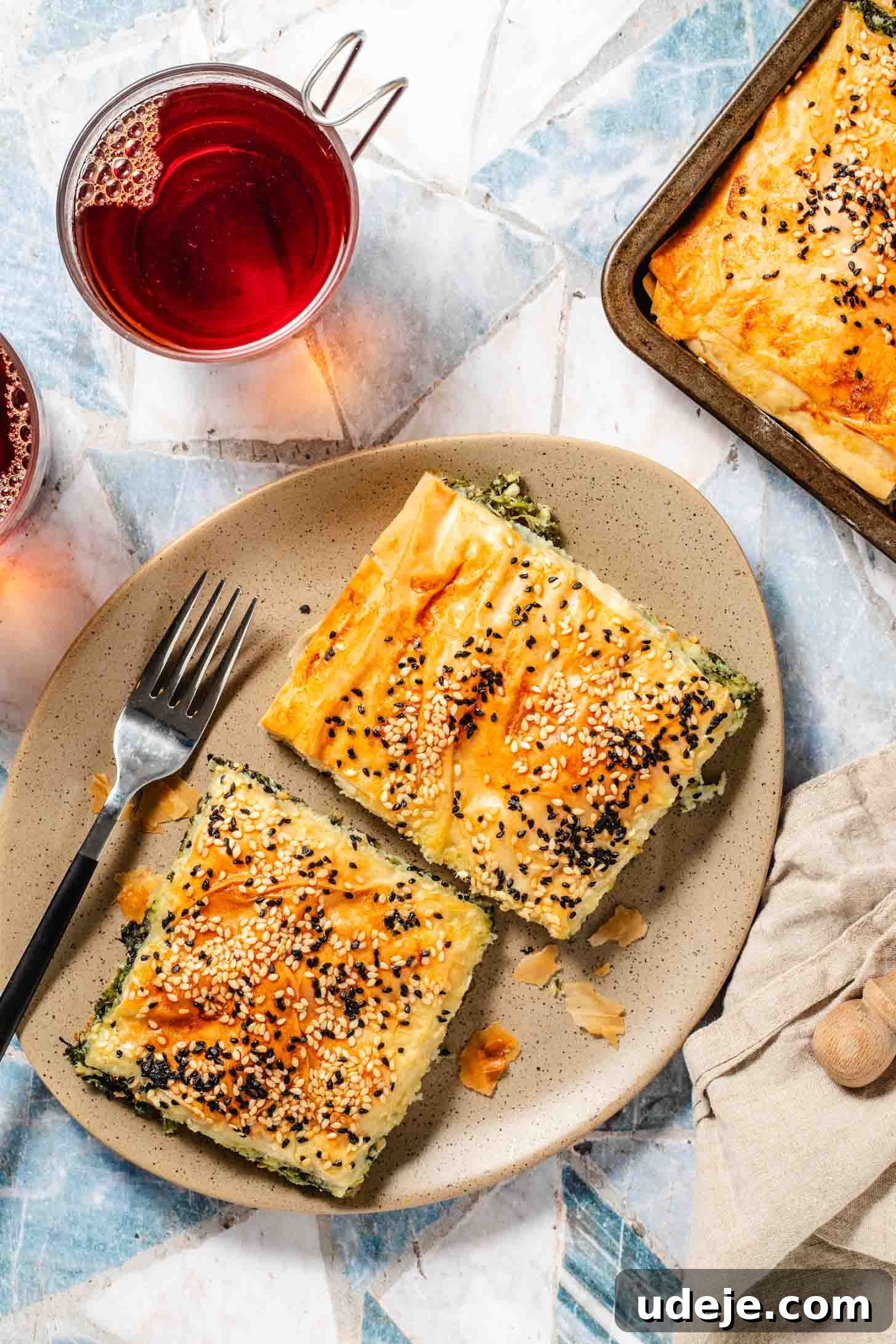 Easy Spinach Borek with Frozen Spinach and Phyllo 2 Two square slices of spinach filled pastry with sesame seeds and nigella seeds on top on an oval beige speckled plate with a fork, with a tray of baked pastry at the side, a beige tea towel and two glass cups of black tea at the side. The borek is golden brown and flaky.