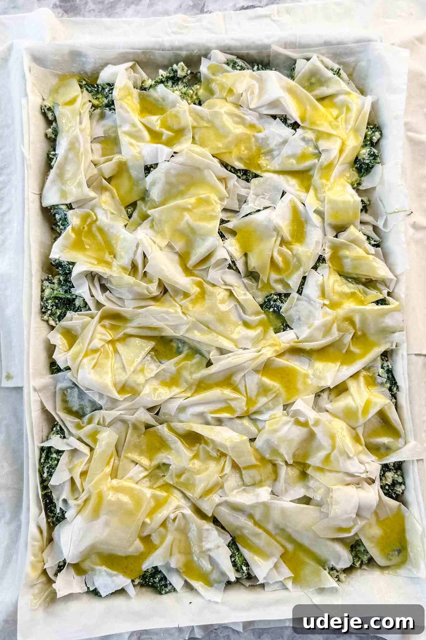 Easy Spinach Borek with Frozen Spinach and Phyllo 11 Phyllo pastry in a sheet pan with a spinach and cheese mixture spread on top with phyllo pastry scrunched on top brushed with oil and melted butter on a marble background. The borek layers are taking shape.