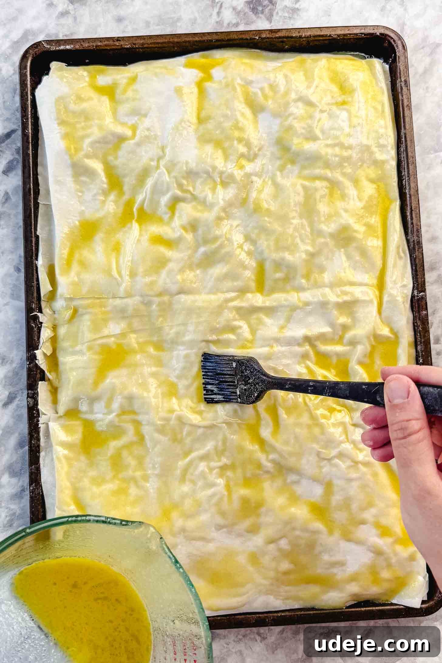 Easy Spinach Borek with Frozen Spinach and Phyllo 14 Folded over phyllo pastry in a sheet pan being brushed with melted butter and olive oil with a black pastry brush and the mixture in a measuring cup on a marble background. The final egg wash is applied.