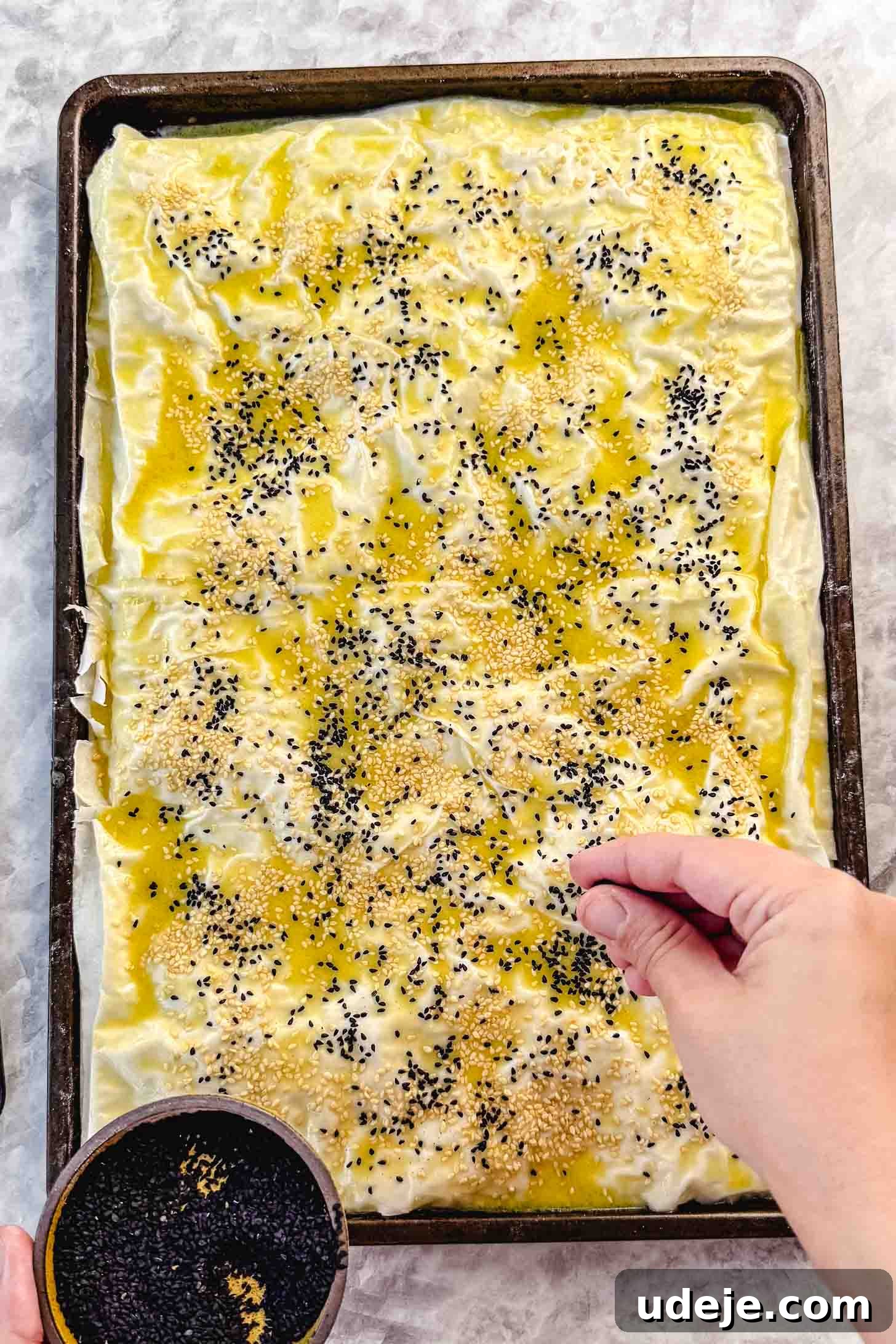 Easy Spinach Borek with Frozen Spinach and Phyllo 15 Folded over phyllo pastry in a sheet pan brushed with melted butter and olive oil and being sprinkled with nigella seeds and sesame seeds on a marble background. The borek is now ready for cutting.