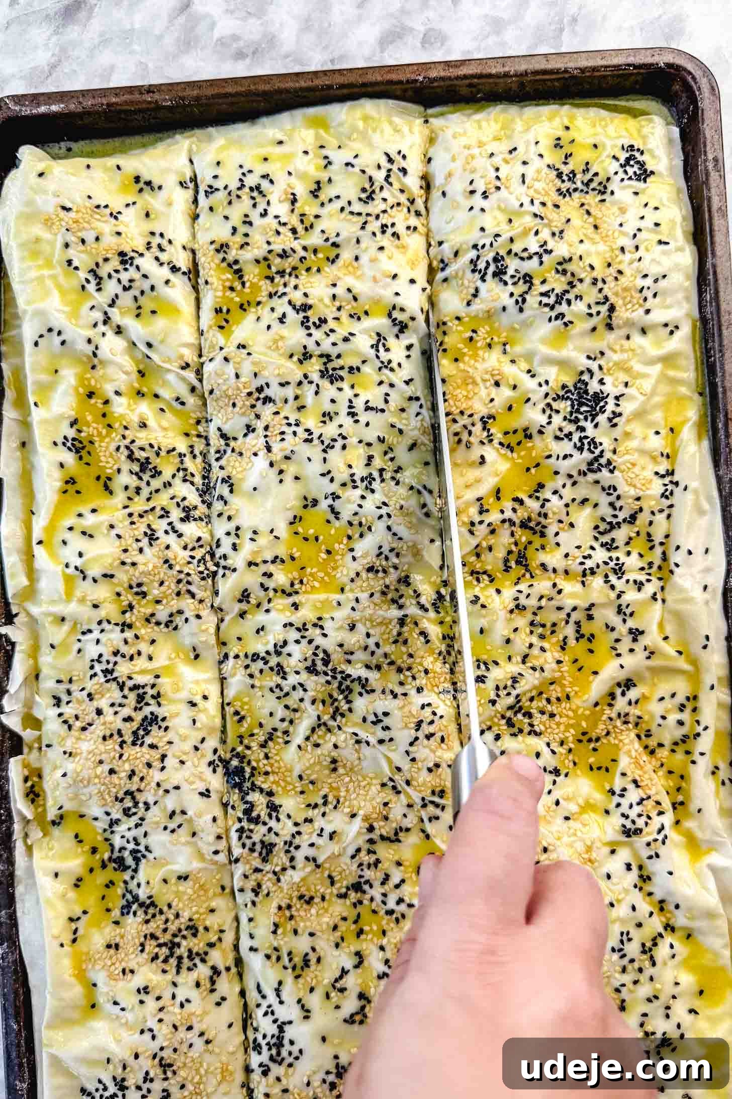 Easy Spinach Borek with Frozen Spinach and Phyllo 16 Folded over phyllo pastry in a sheet pan brushed with melted butter and olive oil and sprinkled with nigella seeds and sesame seeds being cut into squares on a marble background. The borek is being carefully pre-cut.