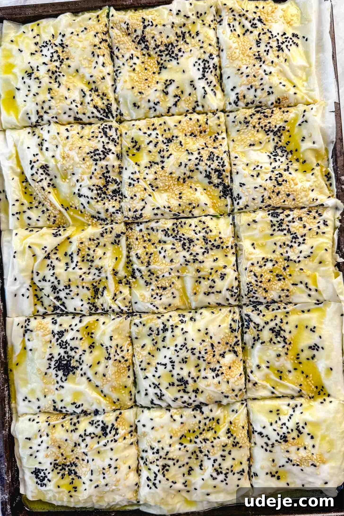 Easy Spinach Borek with Frozen Spinach and Phyllo 17 Folded over phyllo pastry in a sheet pan brushed with melted butter and olive oil and sprinkled with nigella seeds and sesame seeds cut into squares on a marble background. The borek is now fully cut and ready for baking.