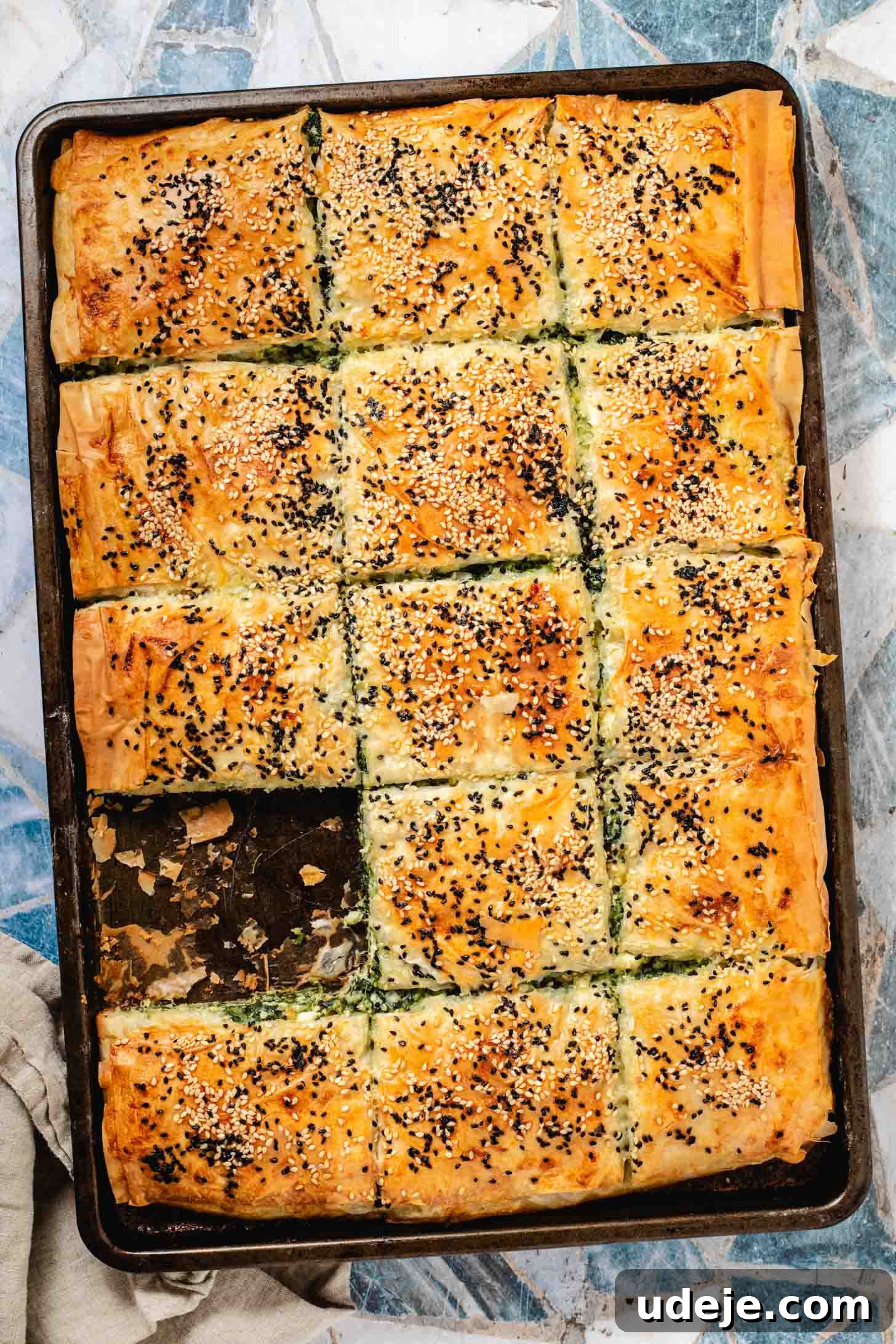 Easy Spinach Borek with Frozen Spinach and Phyllo 18 Spinach filled pastry in a rectangular sheet pan cut into squares with sesame seeds and nigella seeds on top, with one slice missing, a beige tea towel at the side on a light blue and white tiled background. The borek looks golden and ready to serve.
