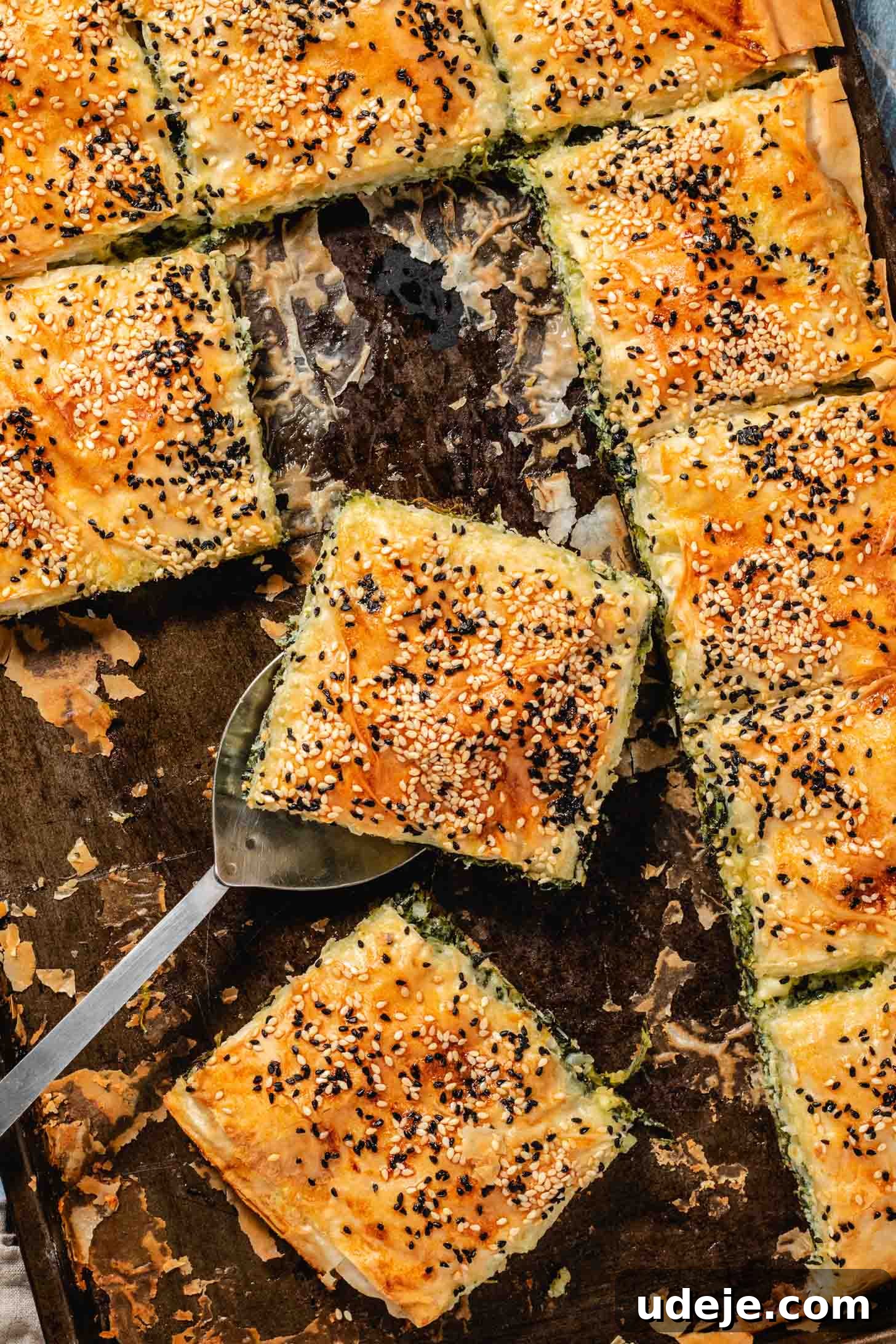 Easy Spinach Borek with Frozen Spinach and Phyllo 19 Spinach filled pastry in a rectangular sheet pan cut into squares with sesame seeds and nigella seeds on top, with one several slices missing and one slice on top of a serving spoon. This highlights the crispy layers and delicious filling.