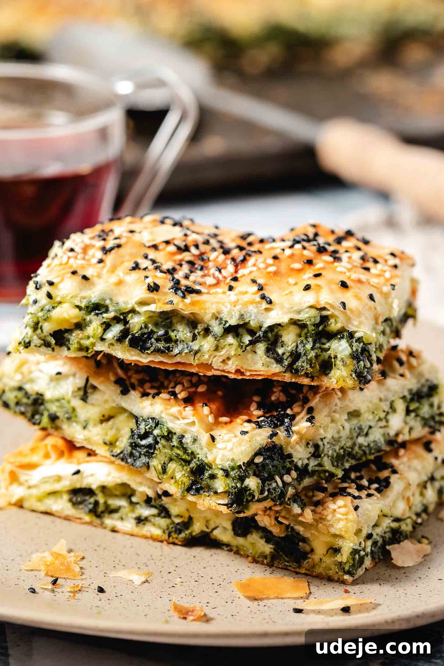Easy Spinach Borek with Frozen Spinach and Phyllo 20 Spinach filled pastry with sesame seeds and nigella seeds on top stacked on top of each other on a beige speckled plate with a glass of black tea behind it along with a tray of baked pastry and a wooden ended serving spoon. This beautiful presentation invites you to try this delicious pastry.