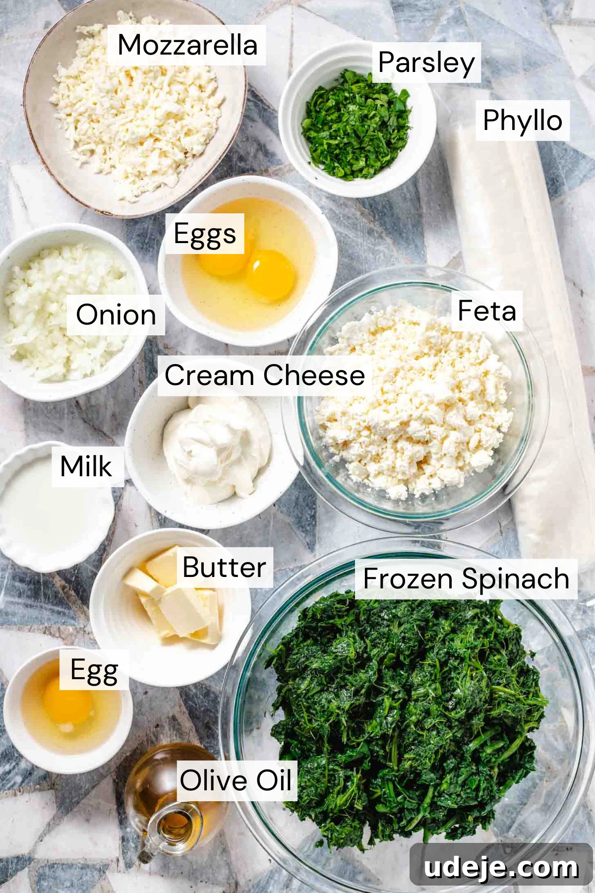 Easy Spinach Borek with Frozen Spinach and Phyllo 3 Ingredient shot with a beige bowl of shredded mozzarella, a small white bowl of chopped parsley, a tube of phyllo dough, a small white bowl with two cracked eggs, a small white bowl of chopped white onion, a glass bowl with feta, a white bowl with cream cheese, a small white bowl with milk, a small white bowl with pats of butter, a small white bowl with one cracked egg, a glass bowl with thawed frozen spinach and a small pitcher of olive oil. All ingredients are neatly arranged on a marble surface.