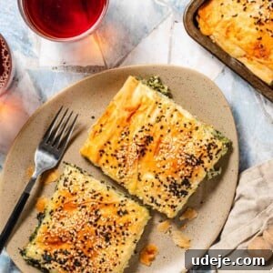 Easy Spinach Borek with Frozen Spinach and Phyllo 21 Two square slices of spinach filled pastry with sesame seeds and nigella seeds on top on an oval beige speckled plate with a fork, with a tray of baked pastry at the side, a beige tea towel and two glass cups of black tea at the side.