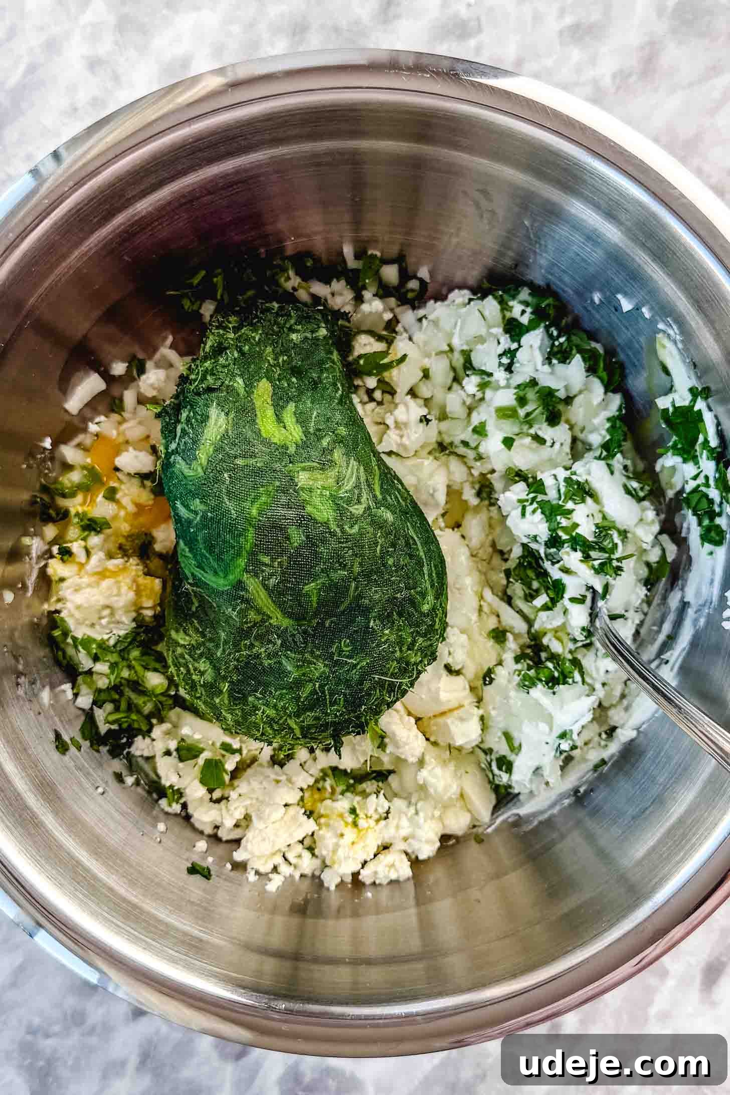 Easy Spinach Borek with Frozen Spinach and Phyllo 4 A metal bowl with crumbled white cheese, frozen spinach, egg and chopped parsley with a metal spoon on a marble background. The ingredients are waiting to be mixed.