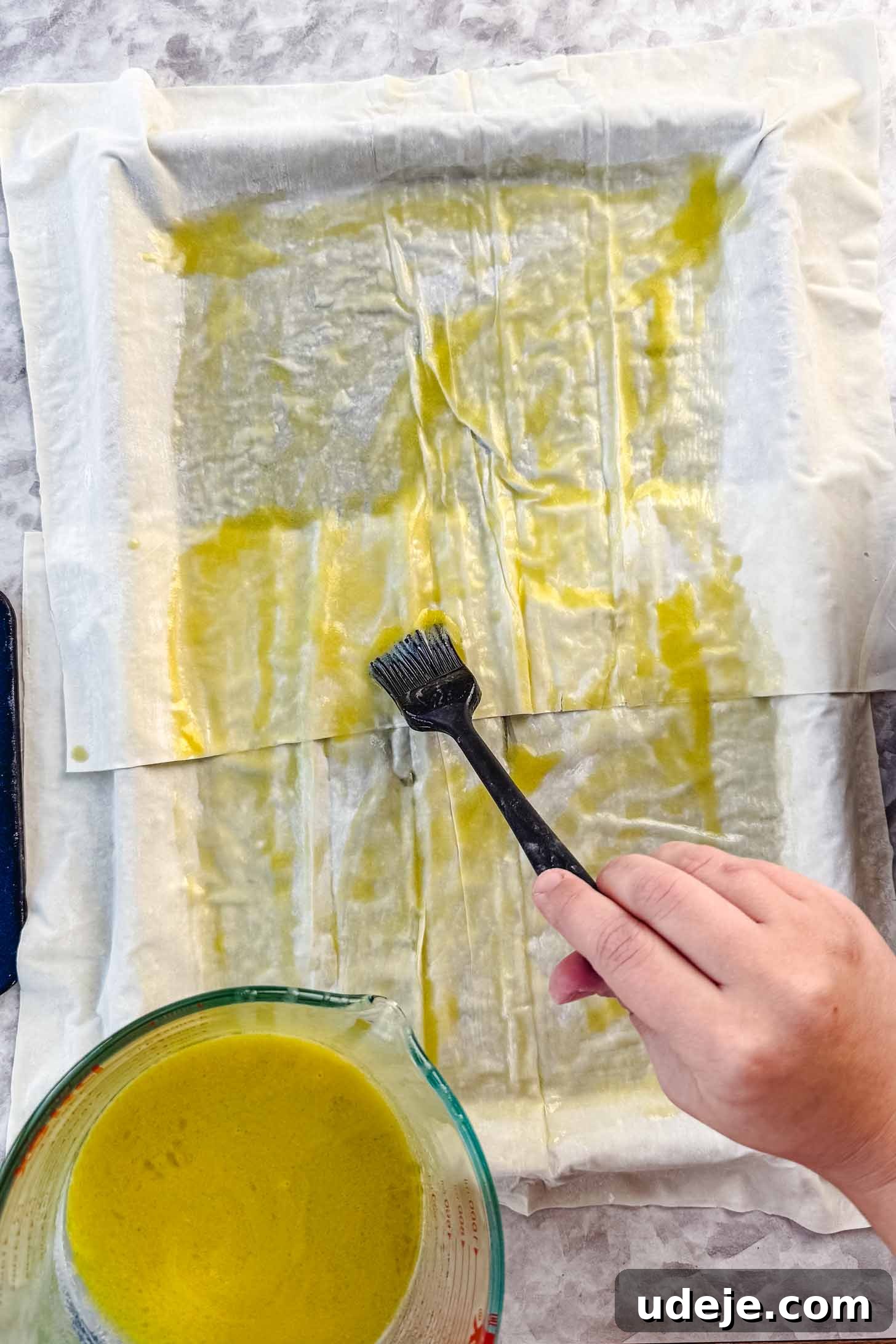 Easy Spinach Borek with Frozen Spinach and Phyllo 7 Phyllo pastry being brushed with butter and olive oil with a black pastry brush on top of the sheet pan with the melted butter and olive oil in a measuring cup on a marble background. The phyllo is being generously coated.