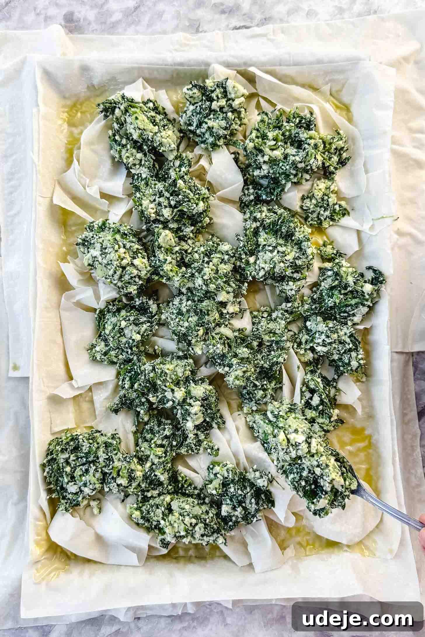 Easy Spinach Borek with Frozen Spinach and Phyllo 9 Phyllo pastry brushed with butter and olive oil on top of the sheet pan with phyllo pastry scrunched up and placed on top with a spinach and cheese mixture spooned on top on a marble background. The filling is being evenly distributed.
