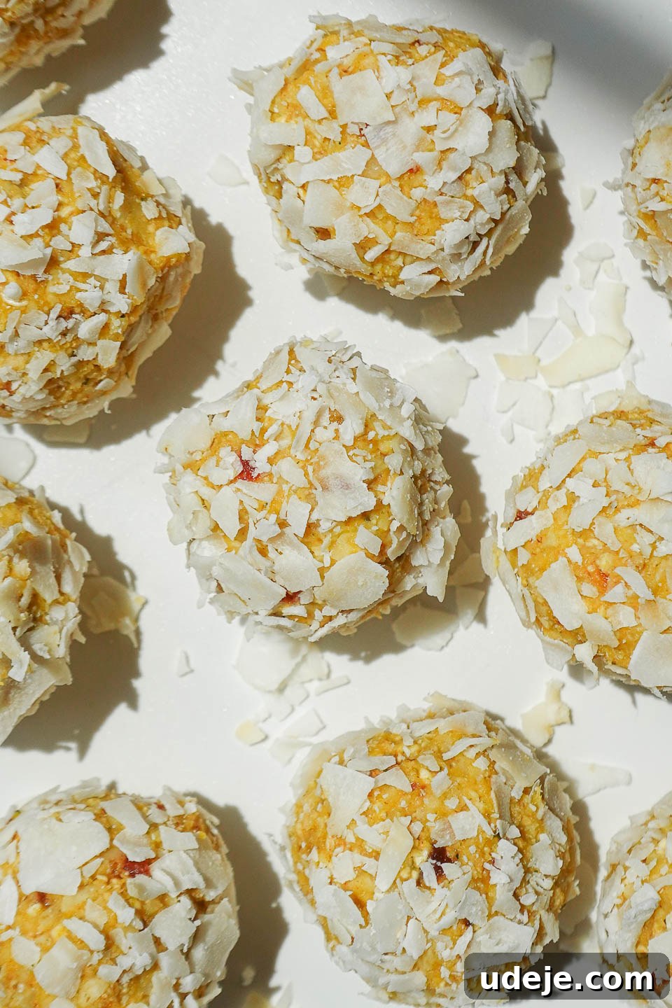 Vegan No-Bake Mango Energy Bites 3 Mango bliss balls being coated in shredded coconut on a plate, showing the texture and preparation process.