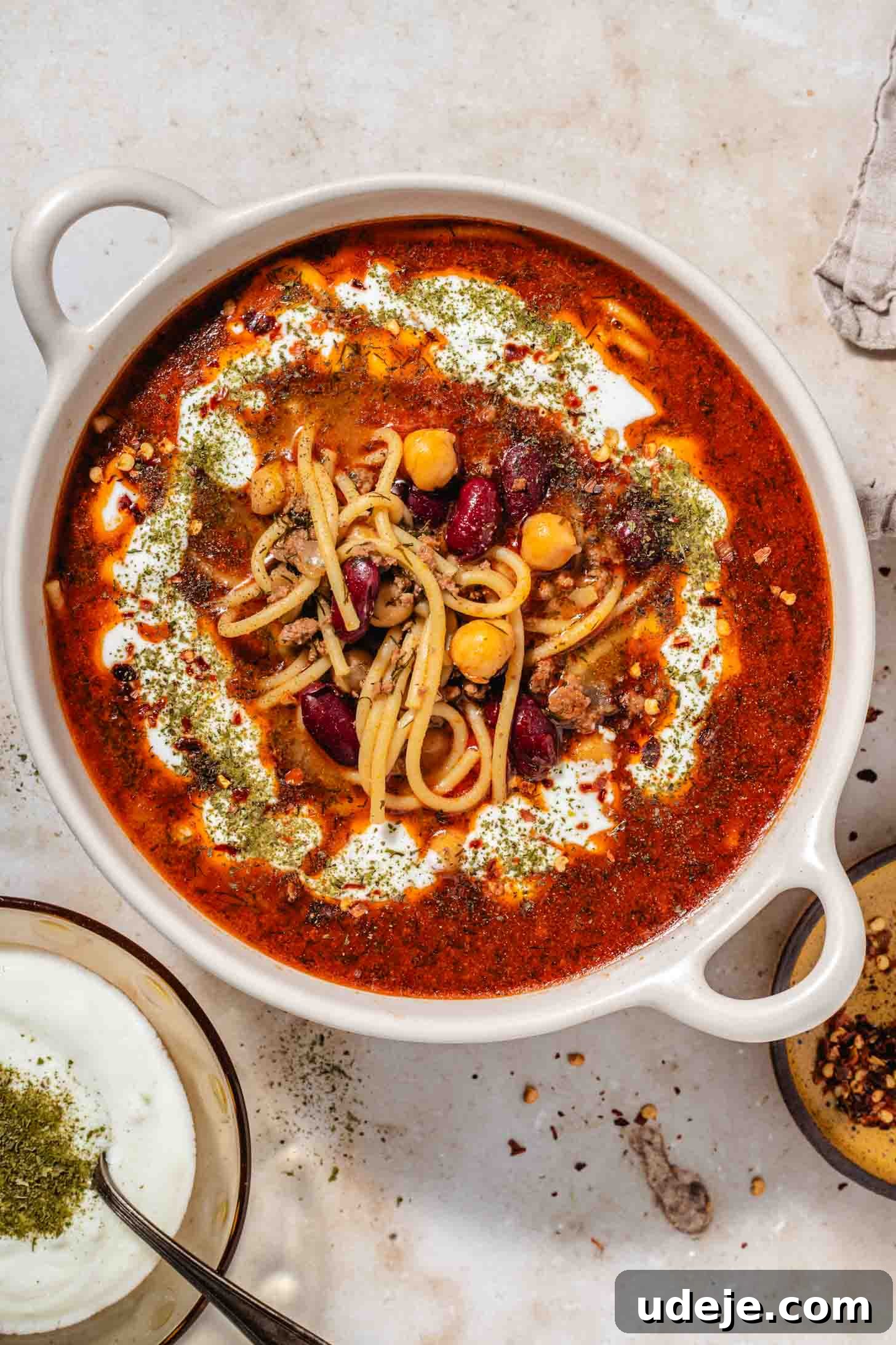 Authentic Afghan Aush Hearty Noodle Delight 2 Creamy bowl with handles filled with a rich red broth, noodles, beans, and a generous drizzle of yogurt and dried mint, served alongside a small bowl of yogurt with dried mint and a spoon, and a small plate with chili flakes, all set on a elegant brown marble background. This image captures the essence of a hearty, flavorful Afghan Aush, a truly divine culinary experience.