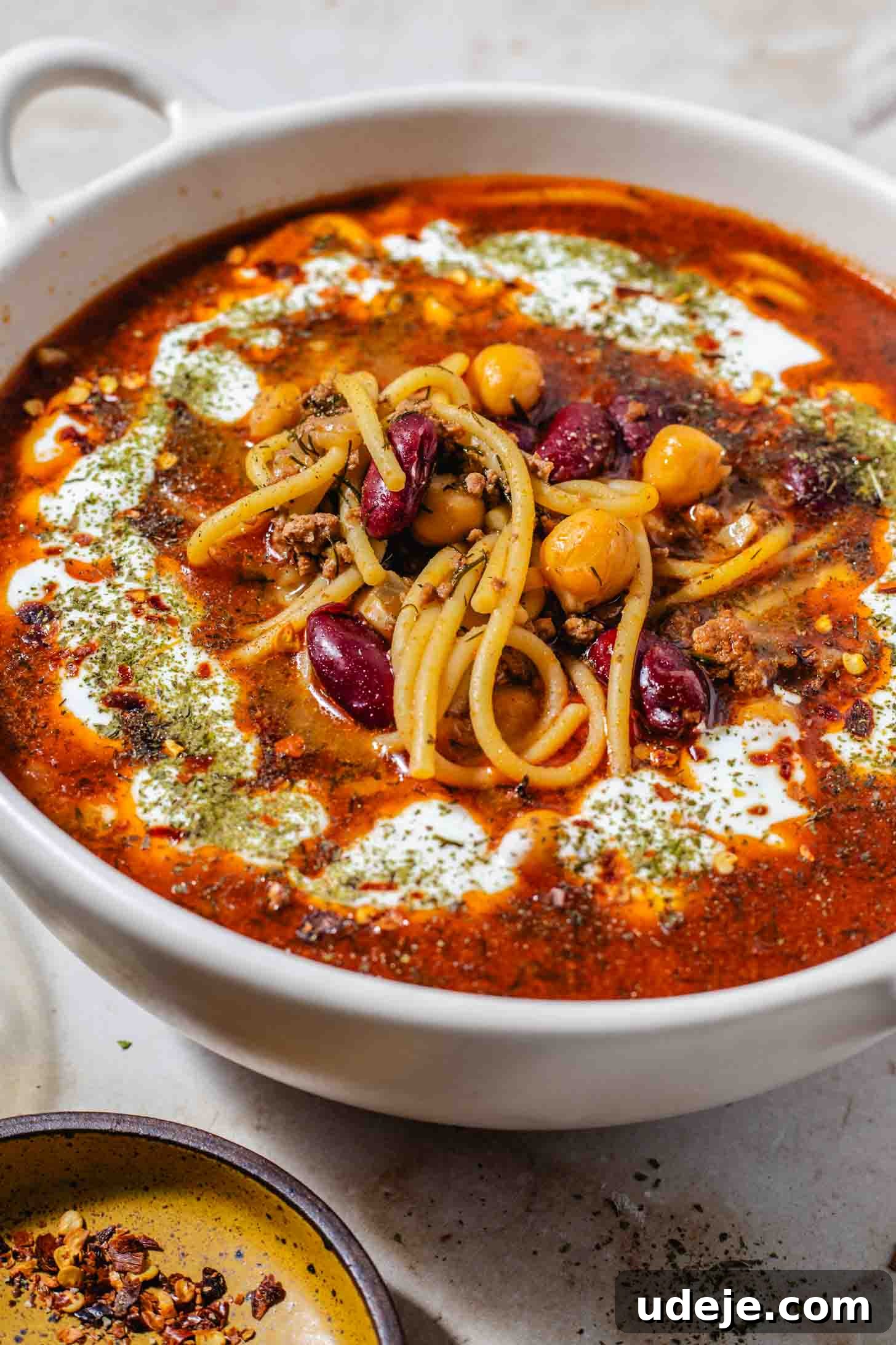 Authentic Afghan Aush Hearty Noodle Delight 12 A creamy bowl with handles, filled with a rich red broth, tender noodles, hearty beans, and a delicate drizzle of yogurt and dried mint. A small plate of vibrant chili flakes sits beside it, all arranged on a rustic brown marble background, showcasing a beautifully garnished Afghan Aush.