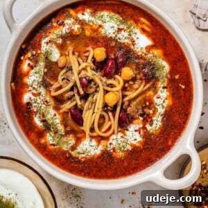 Authentic Afghan Aush Hearty Noodle Delight 13 Creamy bowl with handles filled with a red broth, noodles, beans and a drizzle of yogurt and dried mint with a bowl of yogurt with dried mint and a spoon, a small plate with chilli flakes on a brown marble background.