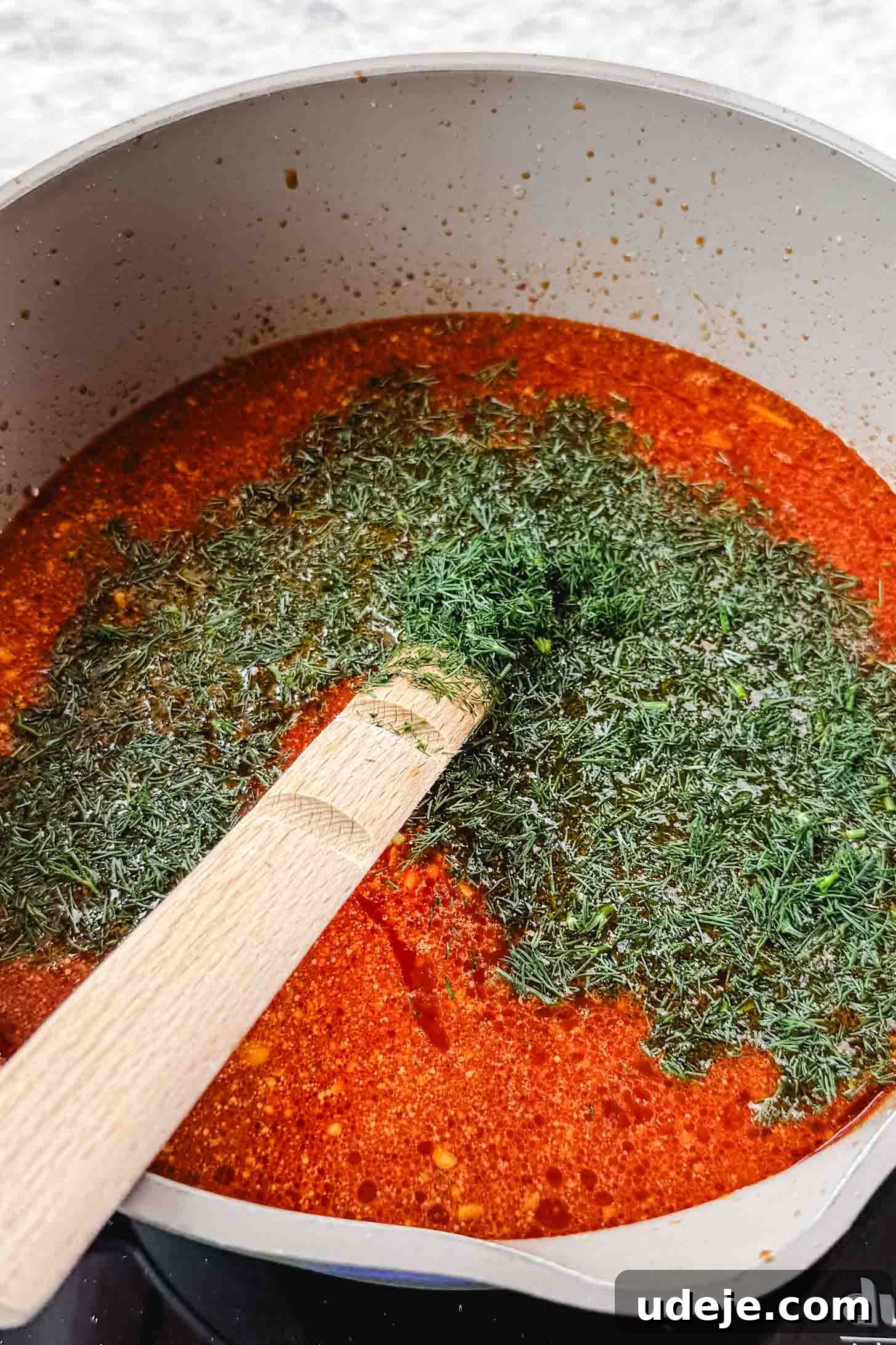 Authentic Afghan Aush Hearty Noodle Delight 9 A vibrant red-colored soup simmering in a cream-colored pot, topped with fresh green dill and a wooden spoon, set against a marble background, showcasing the colorful addition of fresh herbs.