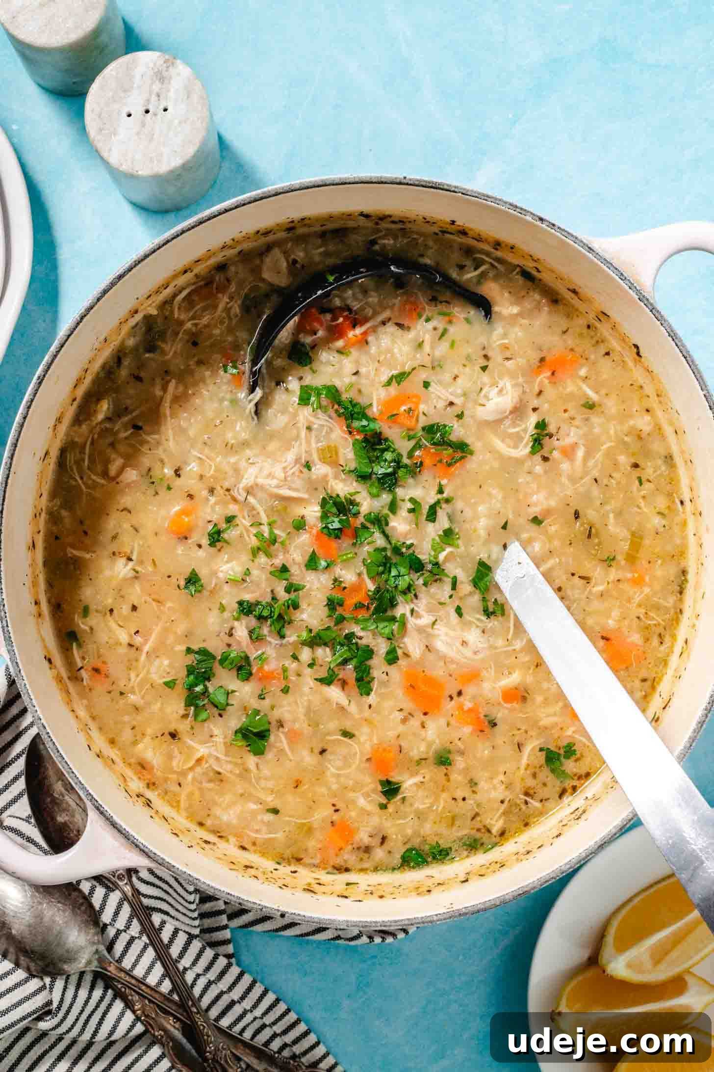 Zesty Lemon Chicken and Rice Soup 2 Cream pot with handles with chicken and rice soup with chopped parsley on top with a metal ladle, a black and white striped towel with 3 spoons, a small plate of lemon wedges and two marble salt and pepper shakers on a light blue background.