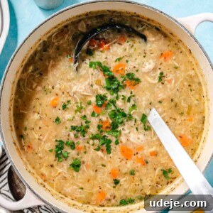 Cream pot with handles with chicken and rice soup with chopped parsley on top with a metal ladle, a black and white striped towel with 3 spoons, a small plate of lemon wedges and two marble salt and pepper shakers on a light blue background.