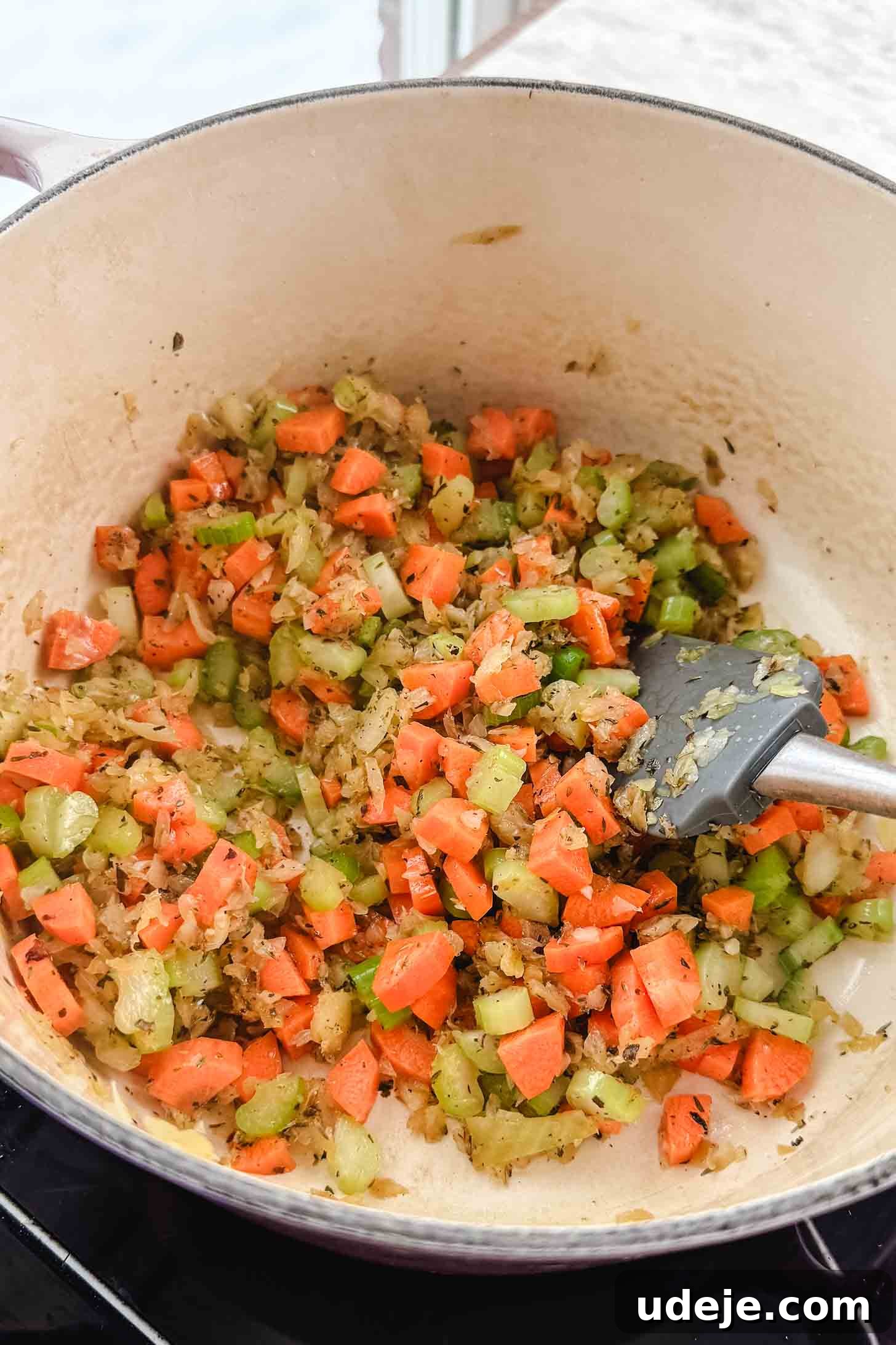 Zesty Lemon Chicken and Rice Soup 4 A cream pot with chopped carrots, onions and celery cooking together with a grey spatula on a marble background.