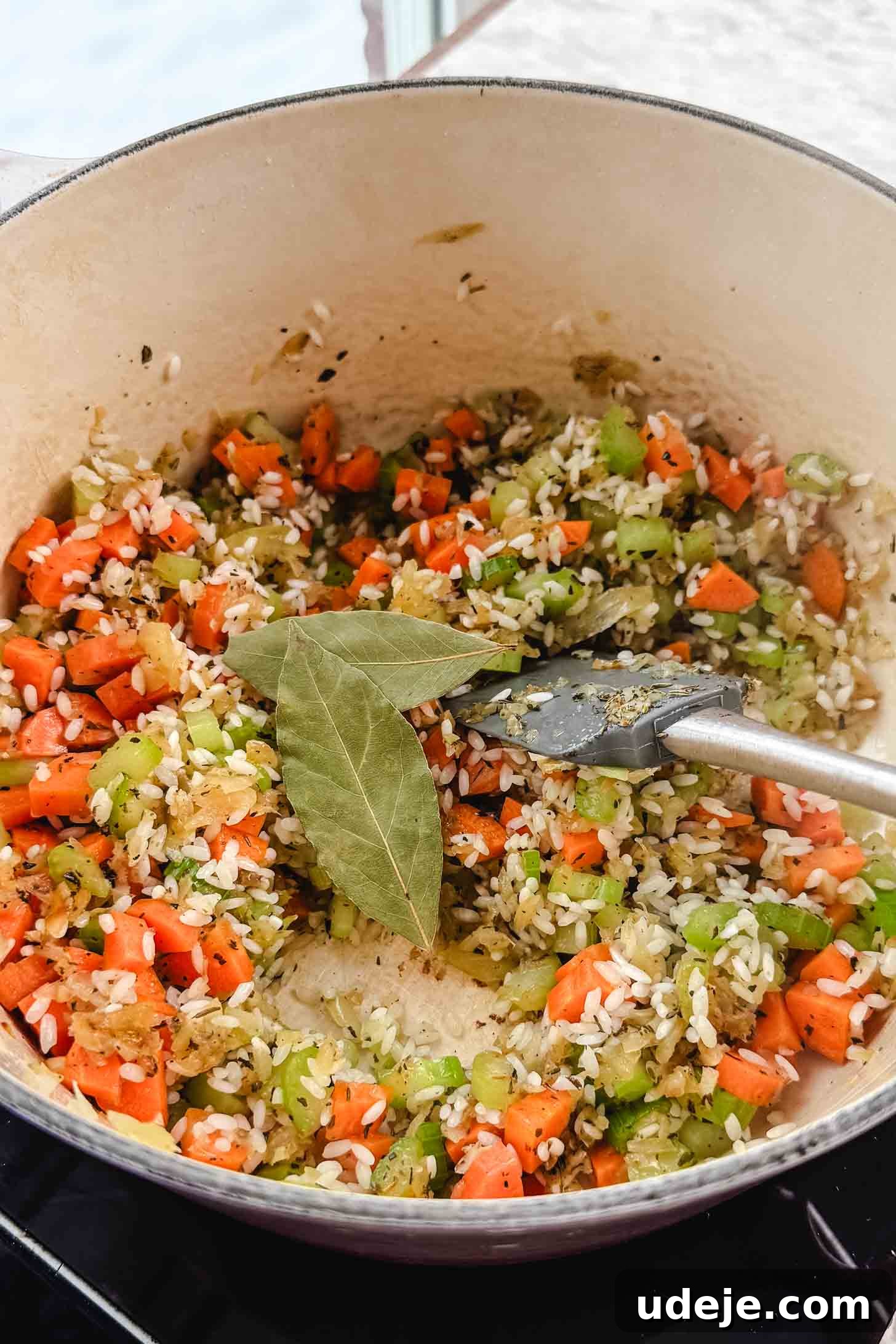 Zesty Lemon Chicken and Rice Soup 5 A cream pot with chopped carrots, onions, bay leaves, uncooked rice and celery cooking together with a grey spatula on a marble background.