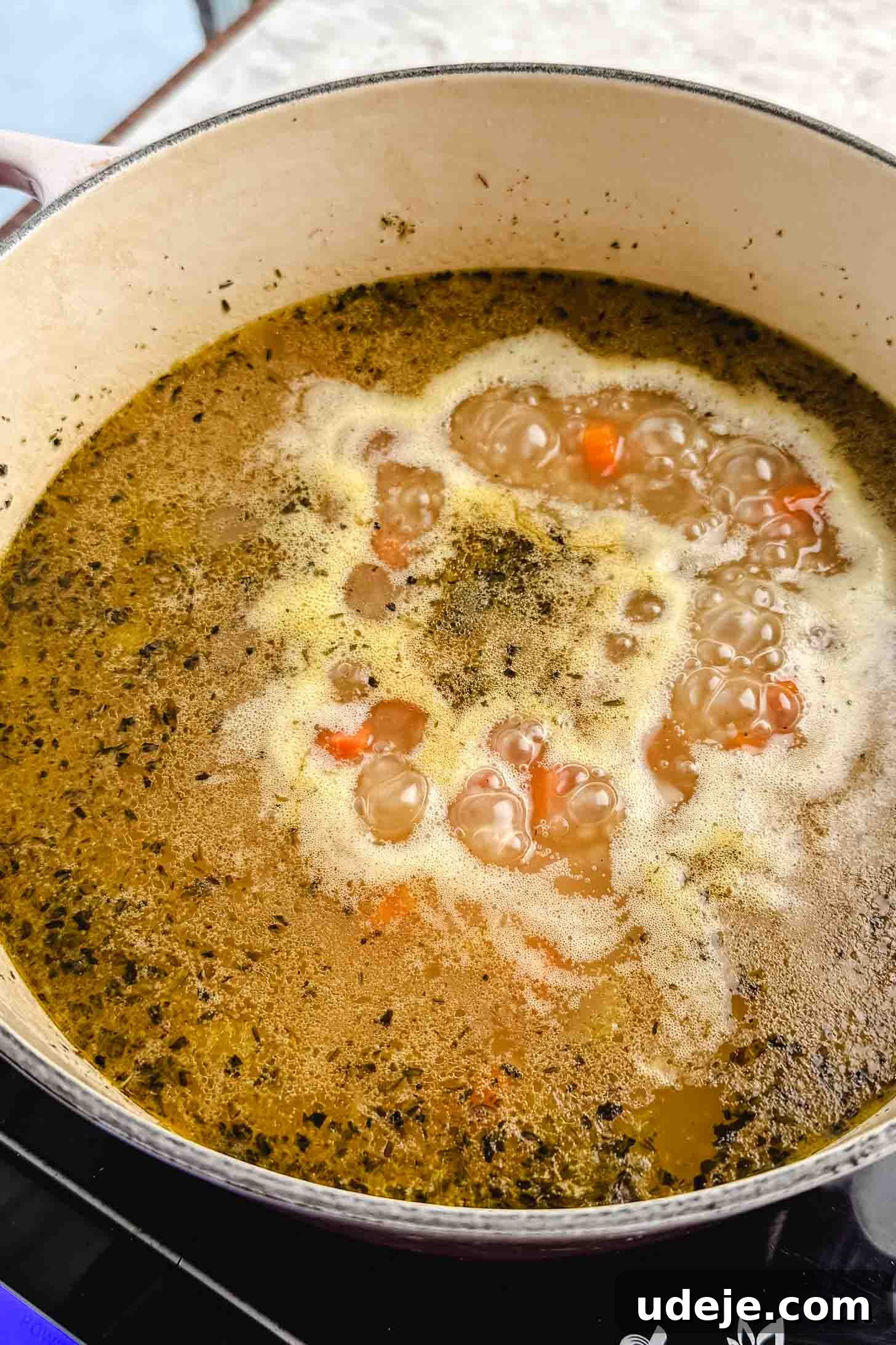 Zesty Lemon Chicken and Rice Soup 6 A cream pot with chopped carrots, onions, bay leaves, uncooked rice, celery and broth cooking together on a marble background.