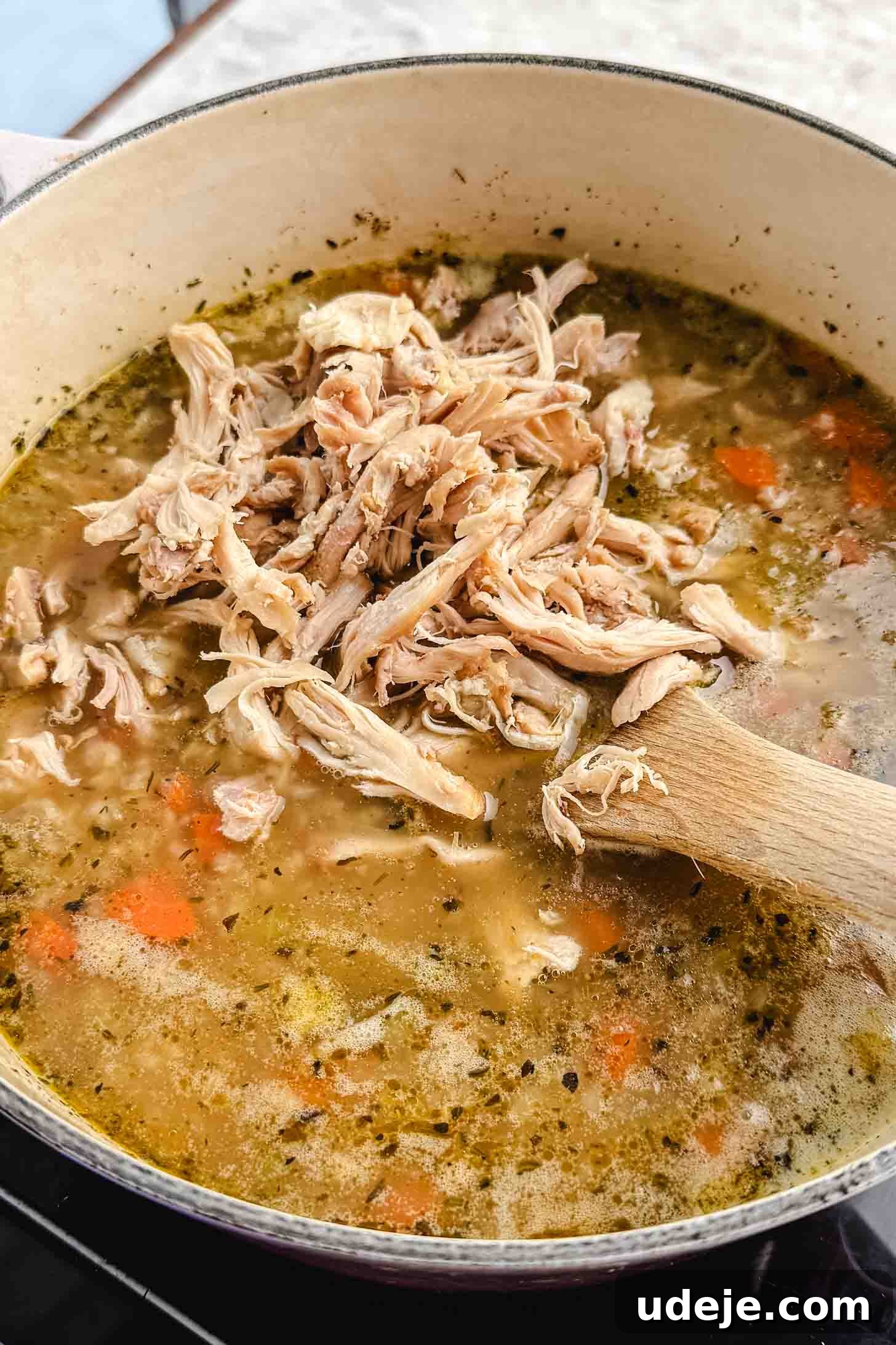 Zesty Lemon Chicken and Rice Soup 7 A cream pot with chopped carrots, onions, bay leaves, uncooked rice, celery, broth and shredded chicken cooking together with a wooden spoon on a marble background.