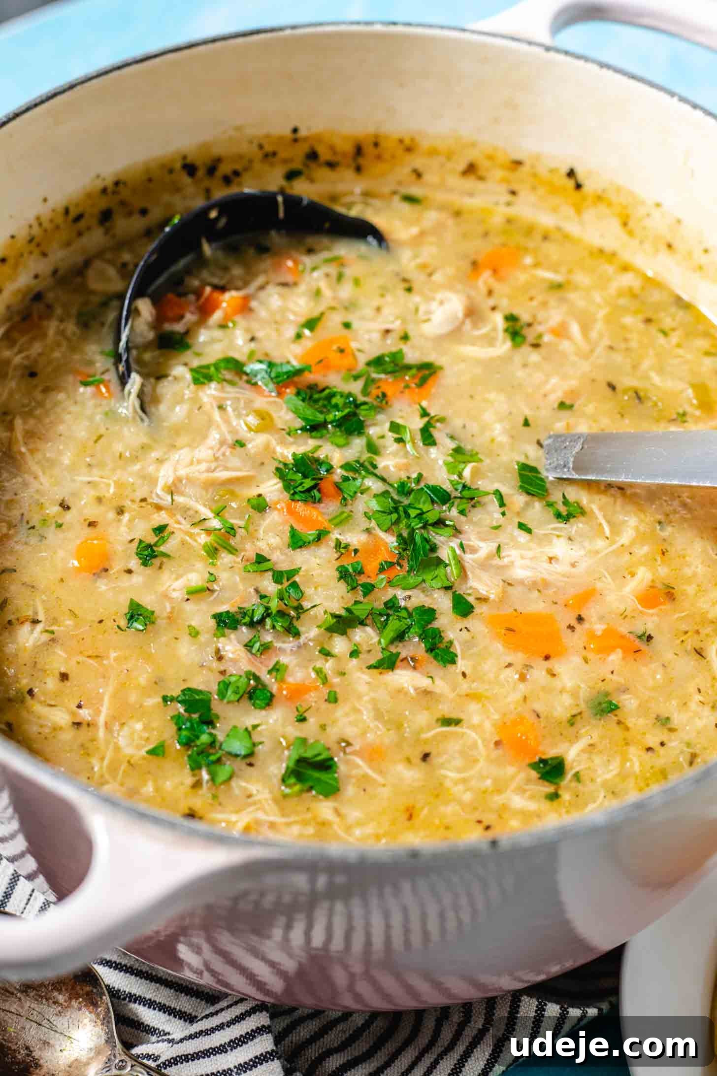 Zesty Lemon Chicken and Rice Soup 8 Cream pot with handles with chicken and rice soup with chopped parsley on top with a metal ladle, a black and white striped towel with a spoon, on a light blue background.