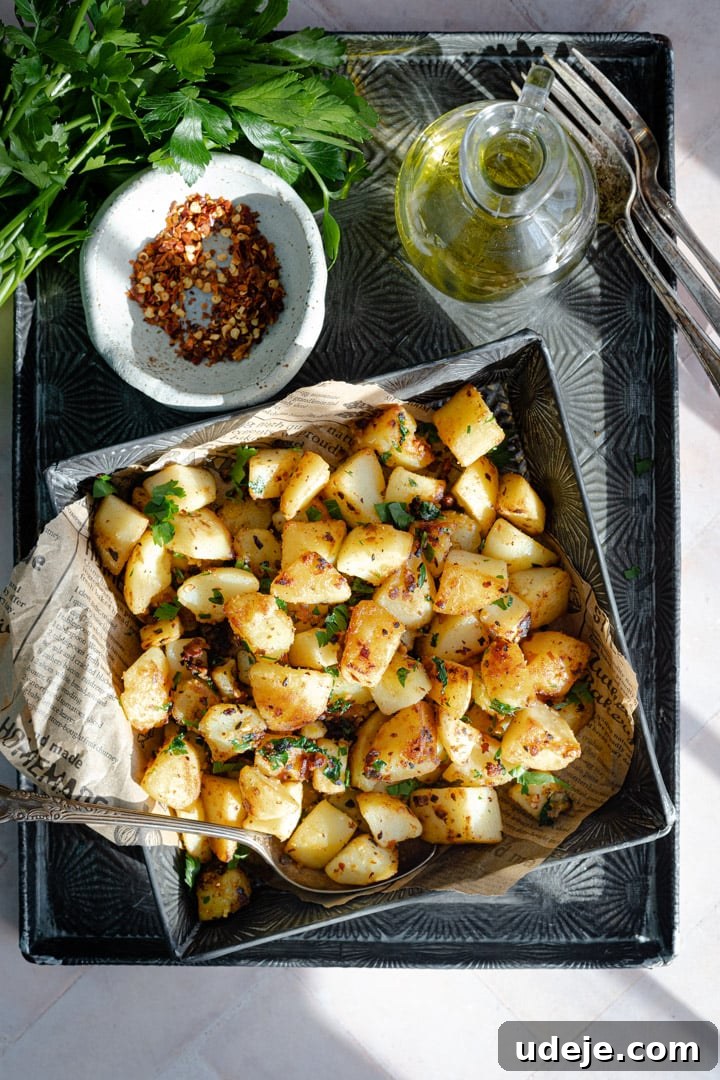 Spicy Golden Crispy Batata Harra 2 cooked crispy potato cubes in a metal tray, lined with parchment paper, garnished with chili flakes and parsley, placed next to a container of olive oil, a bunch of parsley, and a small plate of chili flakes