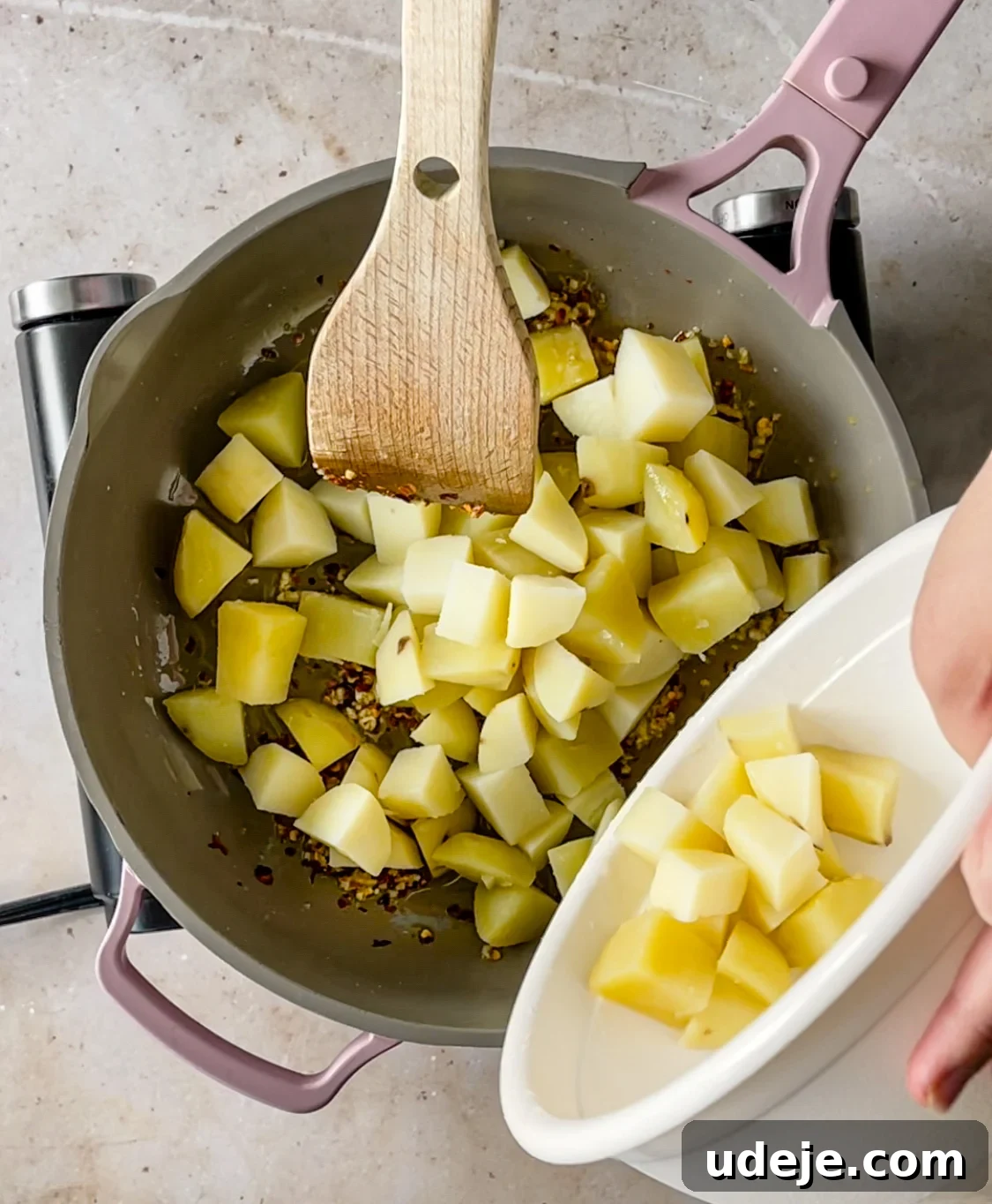 Spicy Golden Crispy Batata Harra 6 olive oil, garlic, and chili flakes in a skillet along with cubed boiled potatoes being cooked using a wooden spoon