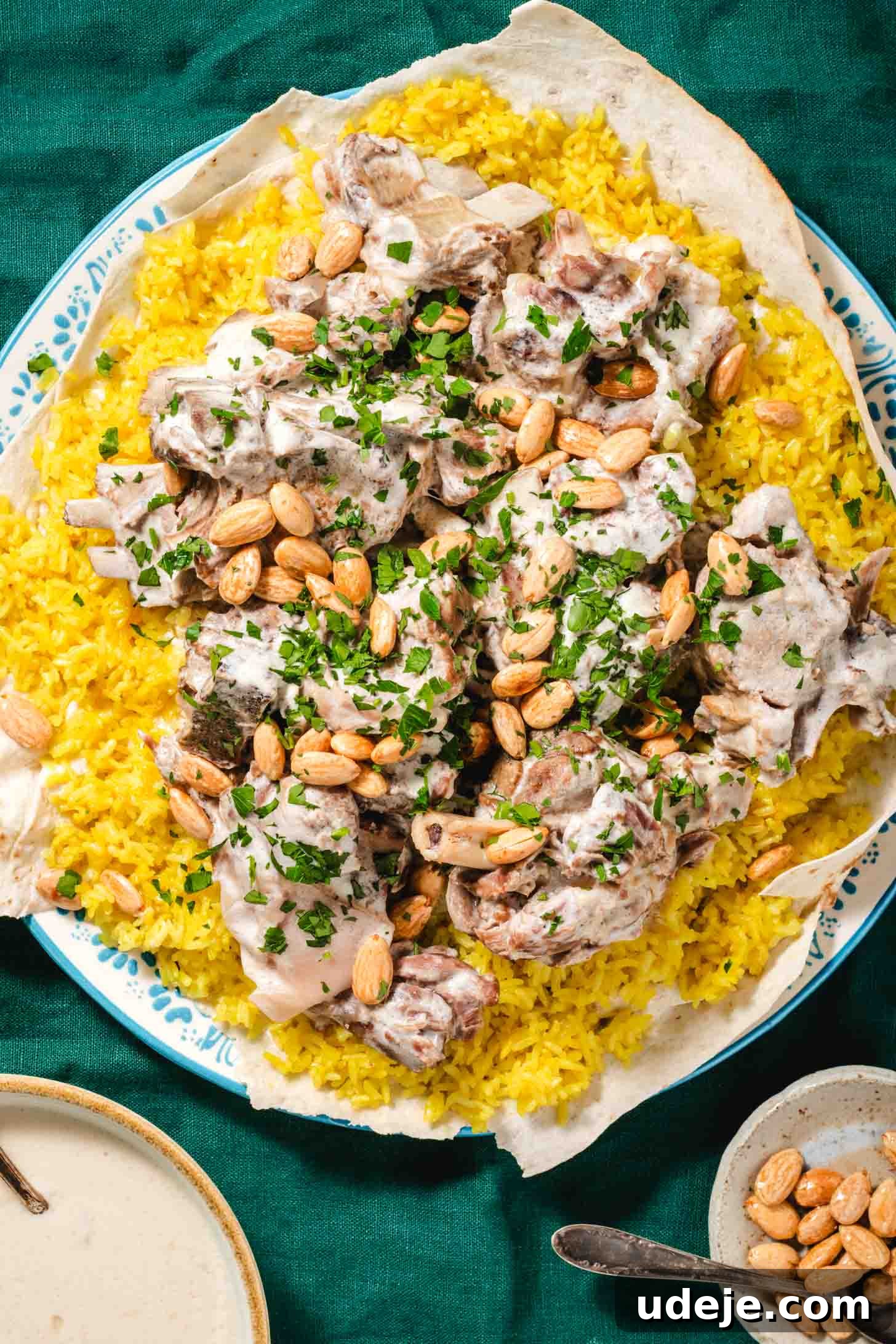 Mansaf: Jordan's Tangy Lamb Masterpiece 2 Cooked lamb shoulder pieces in a white sauce on top of yellow rice that is placed on top of a thin piece of markook on a blue patterned plate. It is garnished with chopped parsley and whole blanched toasted almonds with a beige rimmed bowl of white sauce at the side and a small beige bowl of toasted almonds with a spoon on a dark green tablecloth.