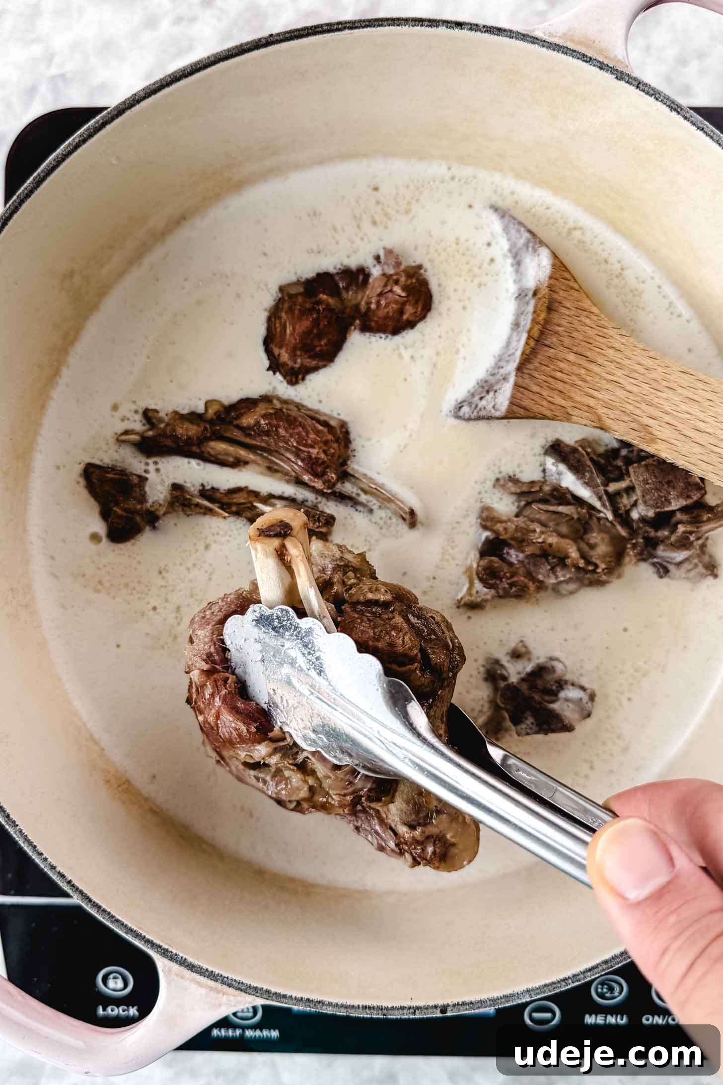 Mansaf: Jordan's Tangy Lamb Masterpiece 11 A beige pot with pink handles, browned and cooked lamb should pieces being placed inside a white liquid with silver tongs and a wooden spoon inside.