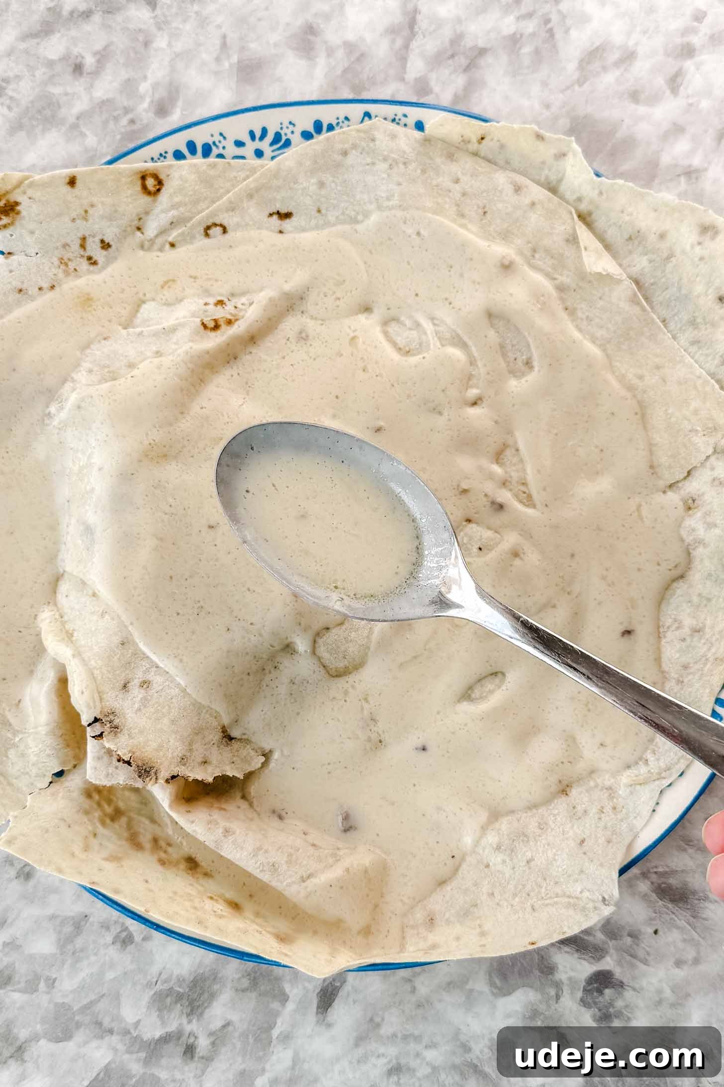 Mansaf: Jordan's Tangy Lamb Masterpiece 12 A blue and white patterned plate with thin pieces of markook bread on top with white liquid being spooned on top with a metal spoon on a marble background.