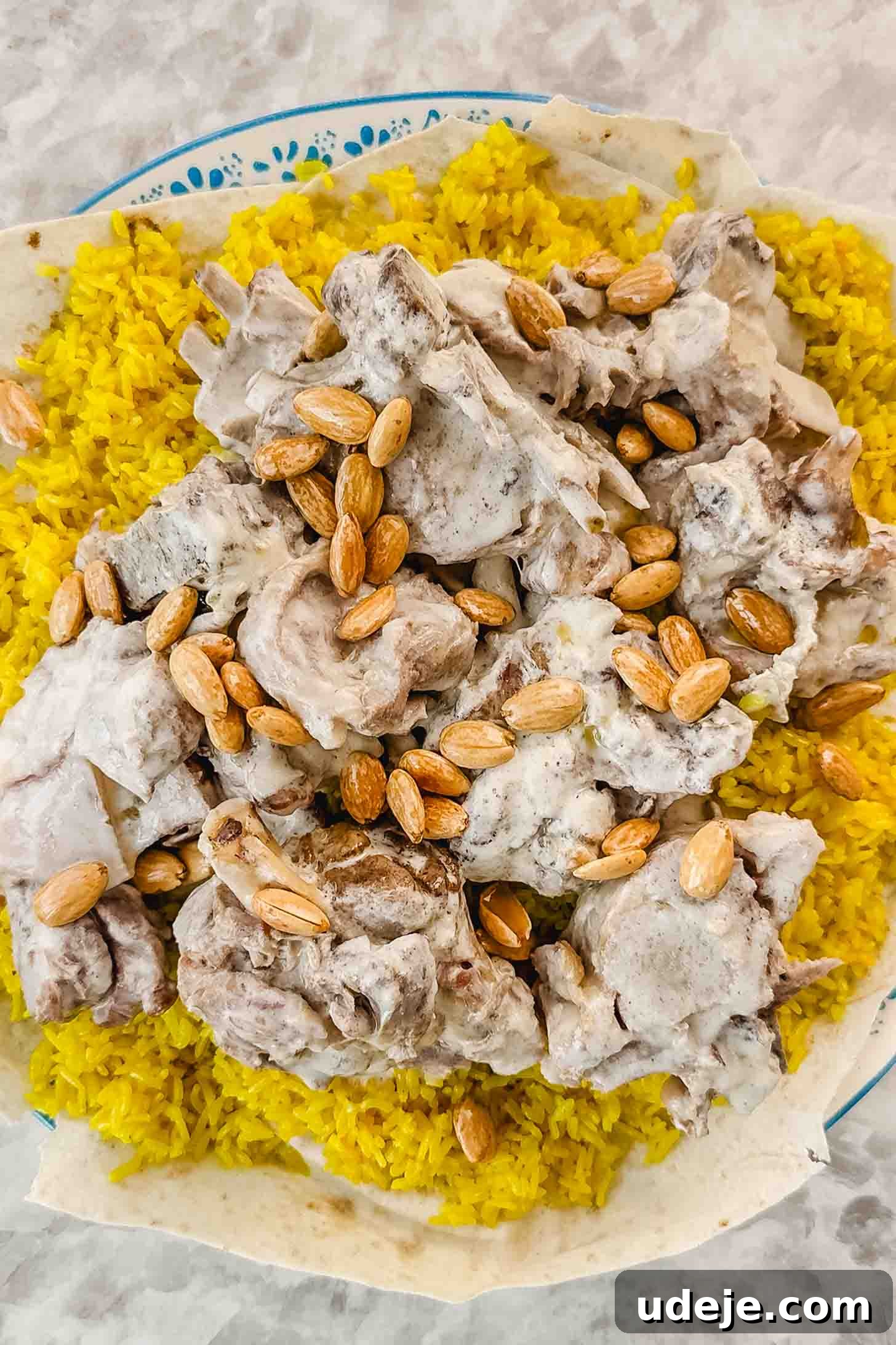 Mansaf: Jordan's Tangy Lamb Masterpiece 14 A blue and white patterned plate with thin pieces of markook bread and yellow rice. Cooked lamb pieces in white sauce and toasted blanched almonds are on top, on a marble background.