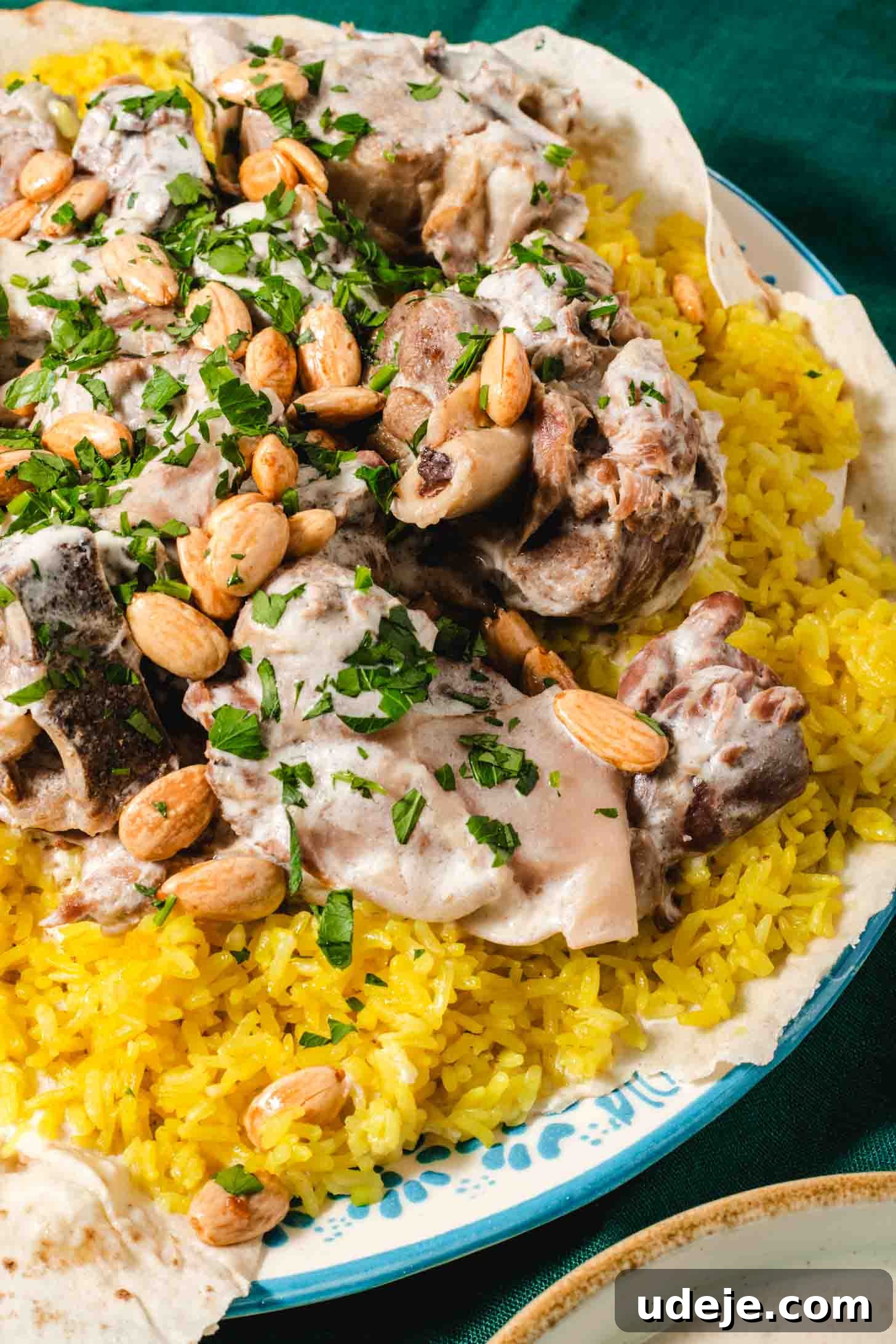 Mansaf: Jordan's Tangy Lamb Masterpiece 15 Cooked lamb shoulder pieces in a white sauce on top of yellow rice that is placed on top of a thin piece of markook on a blue patterned plate. It is garnished with chopped parsley and whole blanched toasted almonds with a beige rimmed bowl of white sauce at the side on a dark green tablecloth.