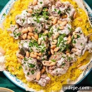 Mansaf: Jordan's Tangy Lamb Masterpiece 16 Cooked lamb shoulder pieces in a white sauce on top of yellow rice that is placed on top of a thin piece of markook on a blue patterned plate. It is garnished with chopped parsley and whole blanched toasted almonds with a beige rimmed bowl of white sauce at the side and a small beige bowl of toasted almonds with a spoon on a dark green tablecloth.