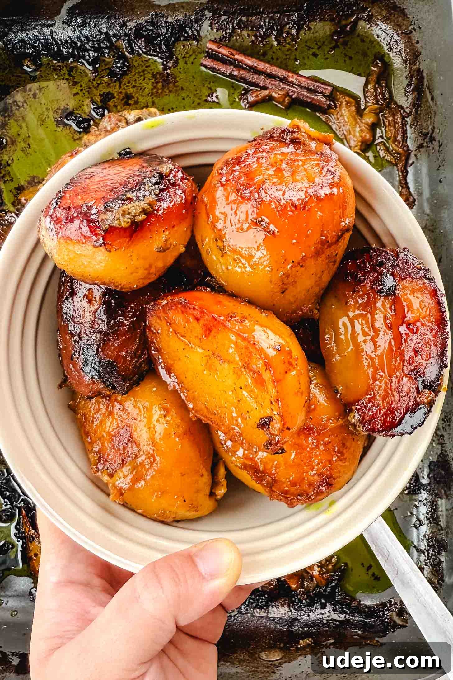 Fall-Apart Tender Lamb Haneeth 11 Roasted halved potatoes in a white hold being held by a hand over the large roasting pan with roasted lamb and onions and a cinnamon stick.