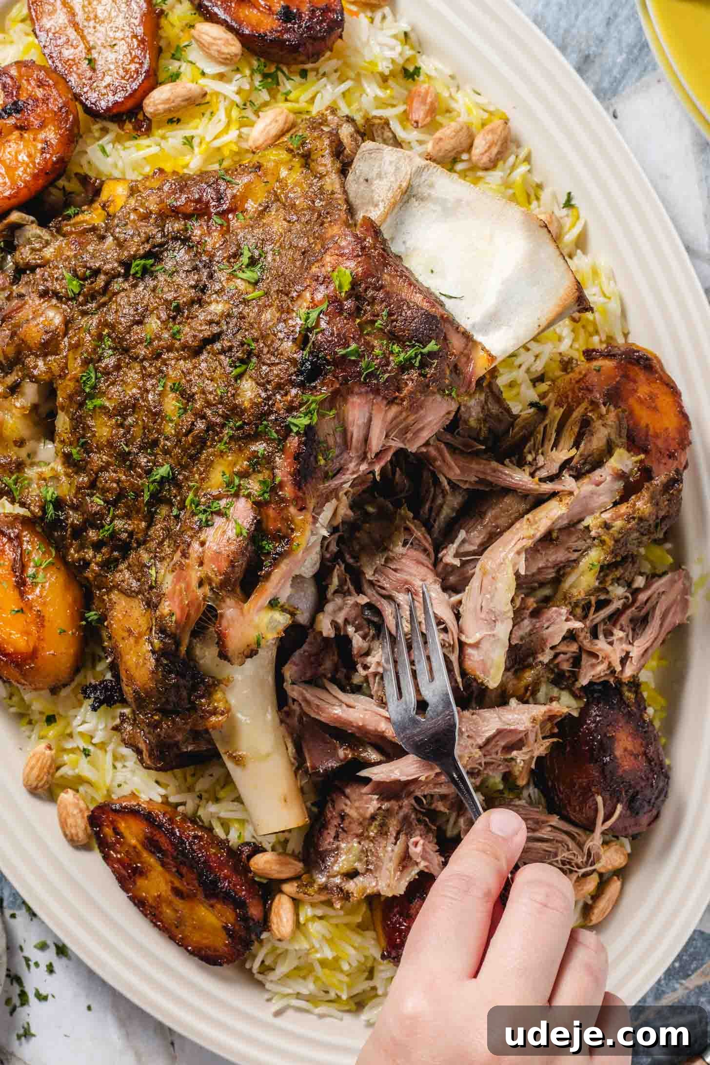 Fall-Apart Tender Lamb Haneeth 13 Baked lamb on a bed of white and yellow rice with roasted potatoes and toasted whole almonds and a sprinkling of parsley with the lamb being shredded with a hand holding a fork on a blue and white tiled background with yellow rimmed plates in the corner.