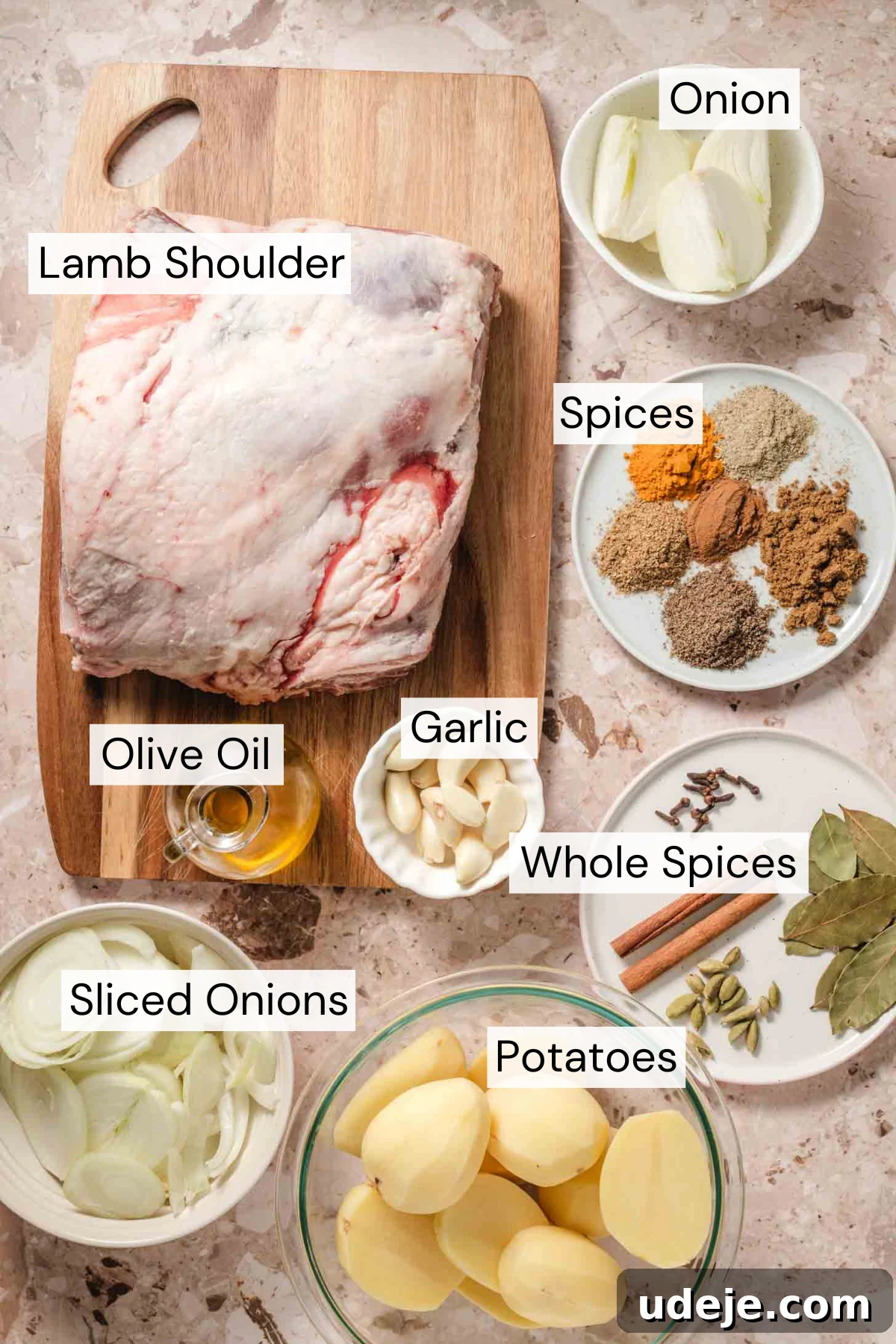 Fall-Apart Tender Lamb Haneeth 3 Ingredient shot with a lamb shoulder on a wooden cutting board, quartered onions in a small white bowl, a white plate with powdered spices, a small bowl with garlic cloves, a small pitcher of olive oil, a white bowl with sliced onion rings, a clear bowl with halved small potatoes and a white plate with whole spices on a beige marble background.