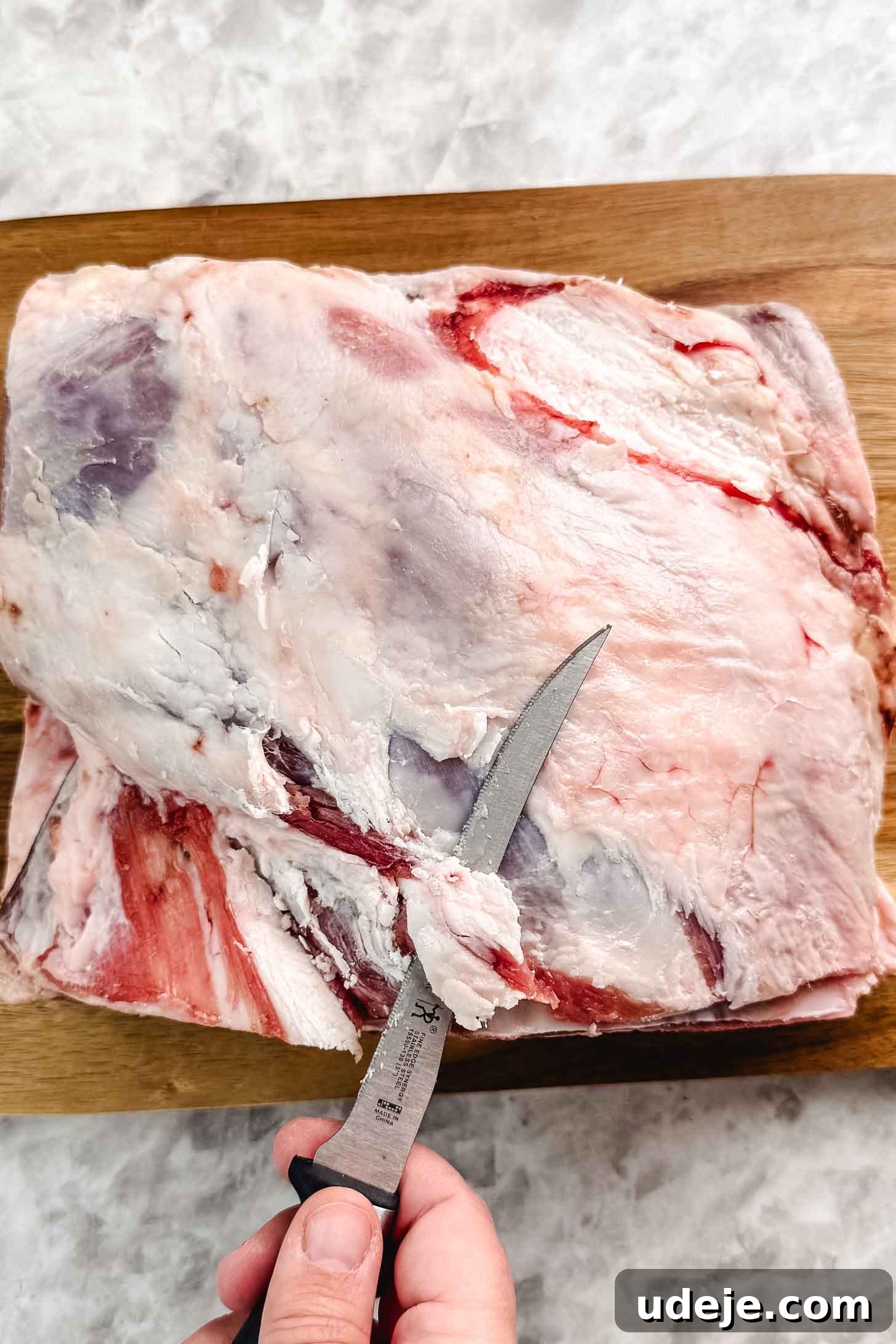 Fall-Apart Tender Lamb Haneeth 6 Lamb shoulder on a wooden cutting board with the fat being trimmed by a knife on a marble background.