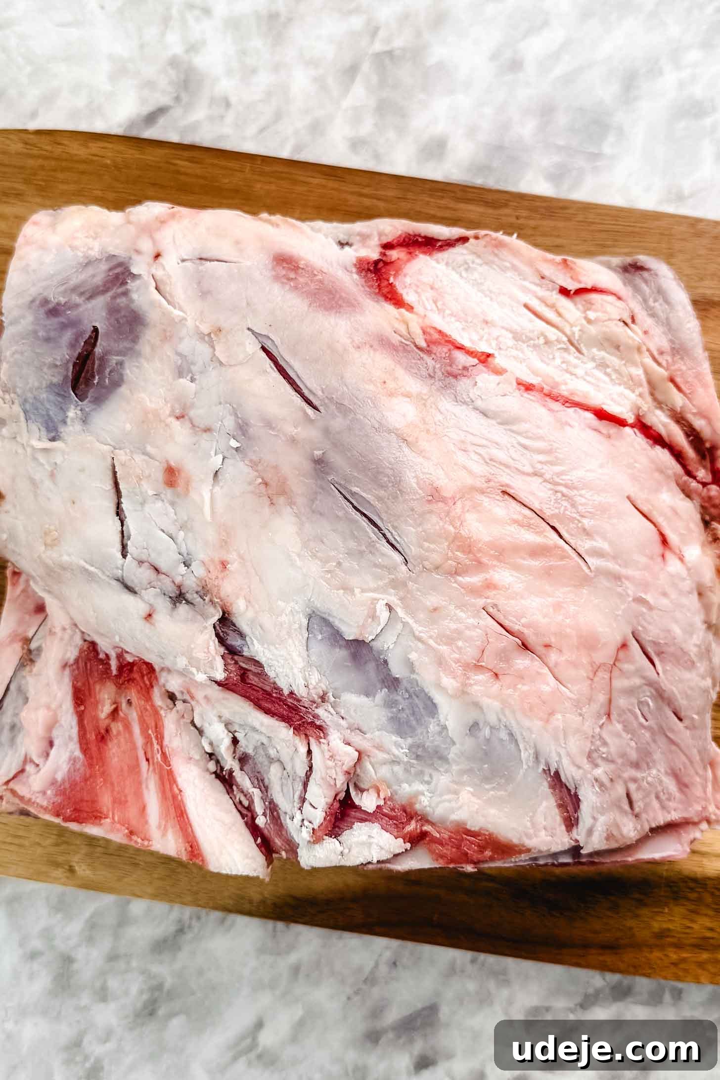 Fall-Apart Tender Lamb Haneeth 7 A lamb shoulder on a wooden cutting board with slits made on the lamb on a marble background.