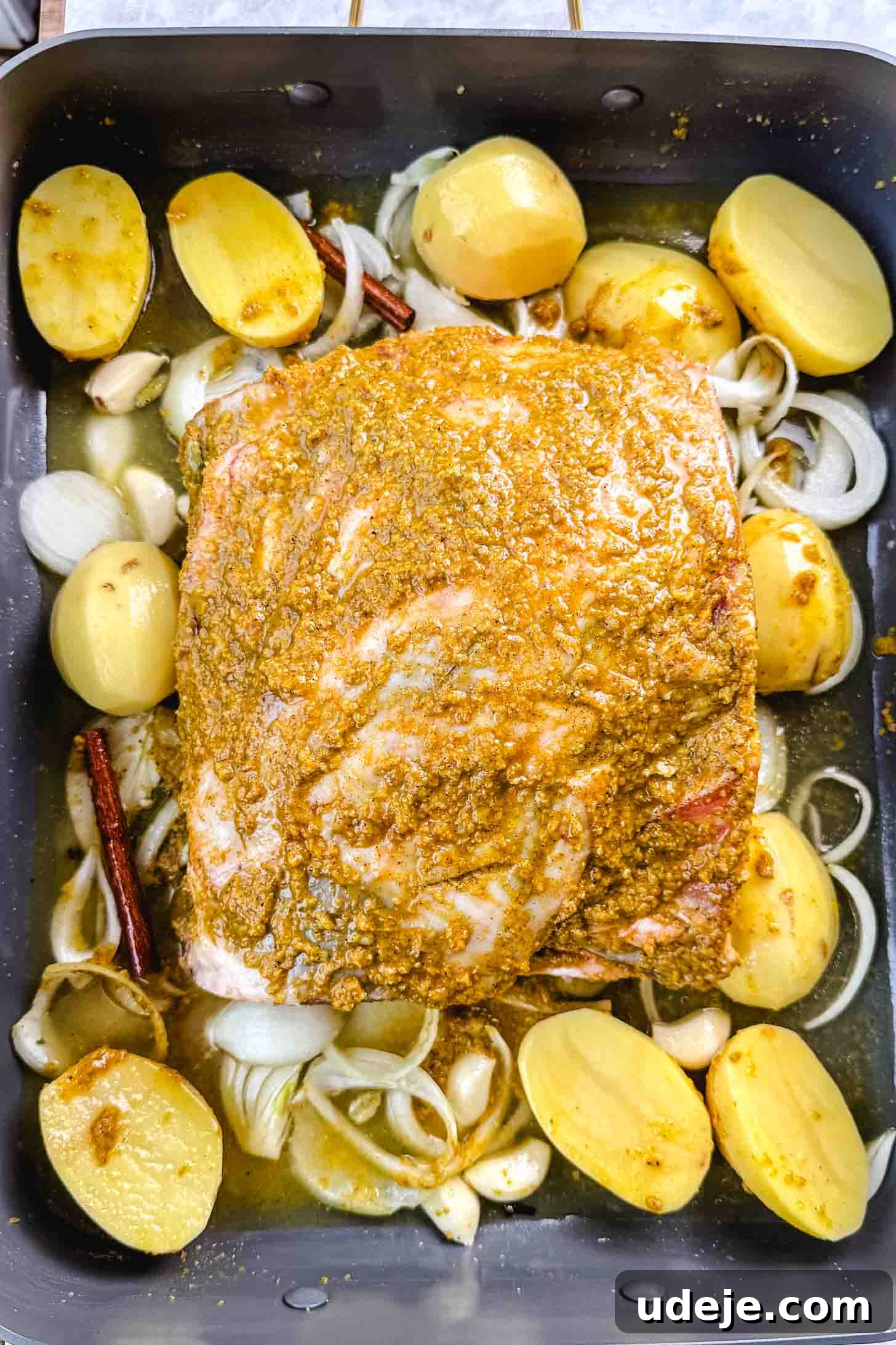 Fall-Apart Tender Lamb Haneeth 9 Large roasting pan with halved potatoes, onion rings, whole garlic cloves, cinnamon sticks, bay leaves and cardamom pods with a drizzle of oil and the lamb shoulder with the brown marinade on top.