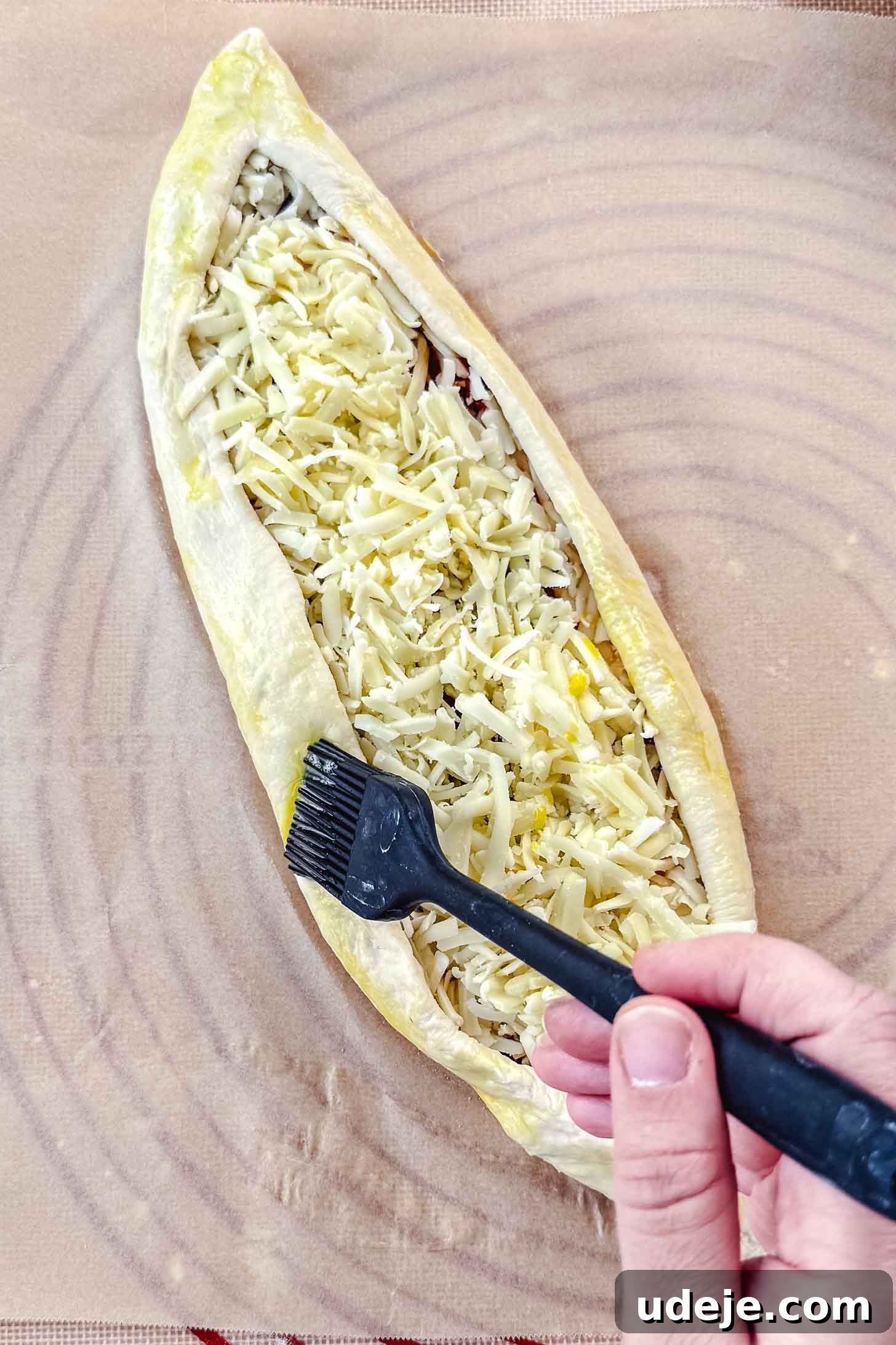 Customizable Homemade Turkish Pide 11 A hand holds a black silicone brush, applying an egg wash to the edges of a boat-shaped dough piece filled with shredded white cheese, resting on parchment paper, to achieve a golden finish.