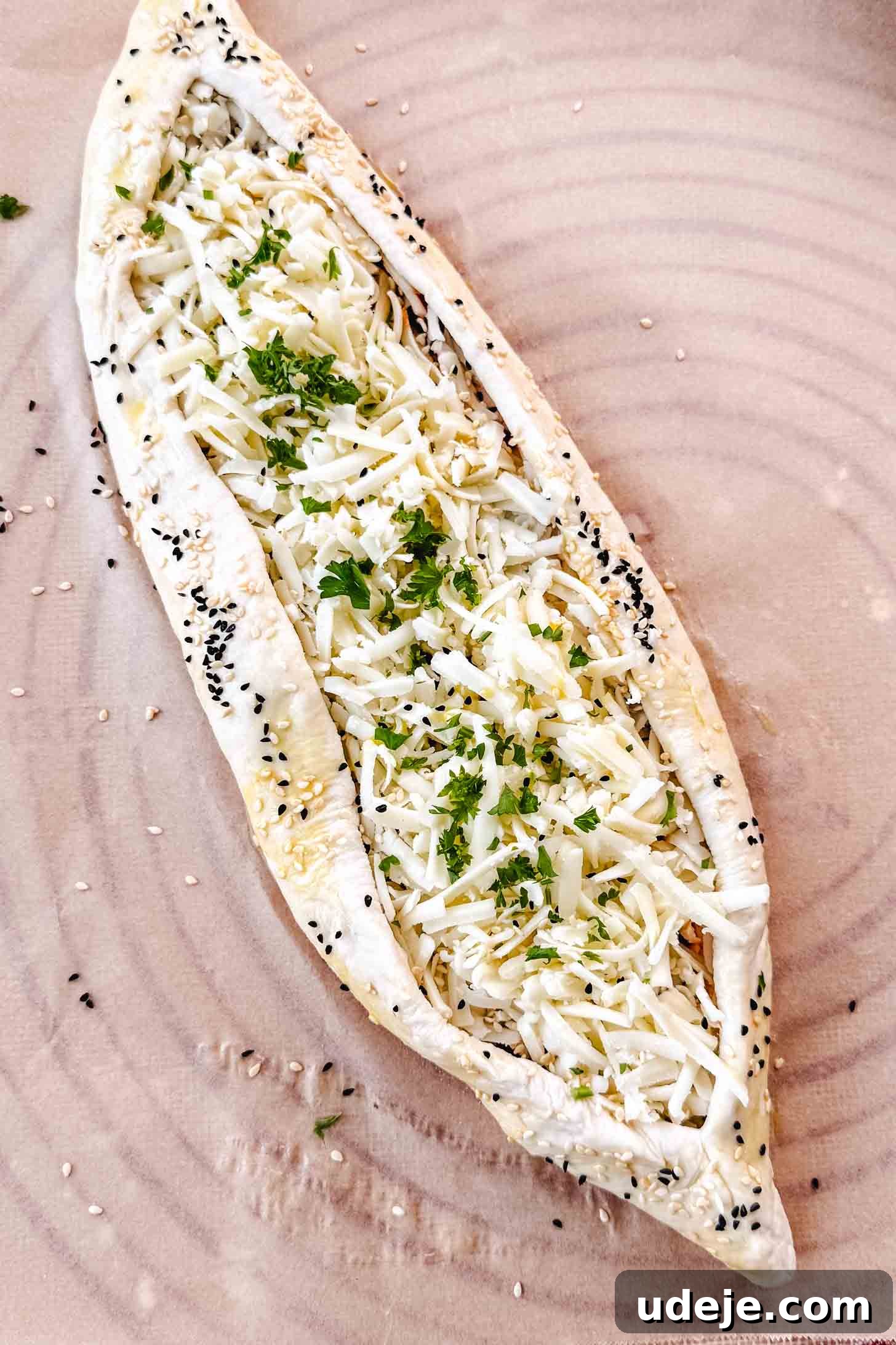 Customizable Homemade Turkish Pide 12 A beautifully formed boat-shaped dough, filled with shredded white cheese and topped with chopped parsley, then generously sprinkled with nigella and sesame seeds, all resting on a piece of parchment paper, ready for baking.
